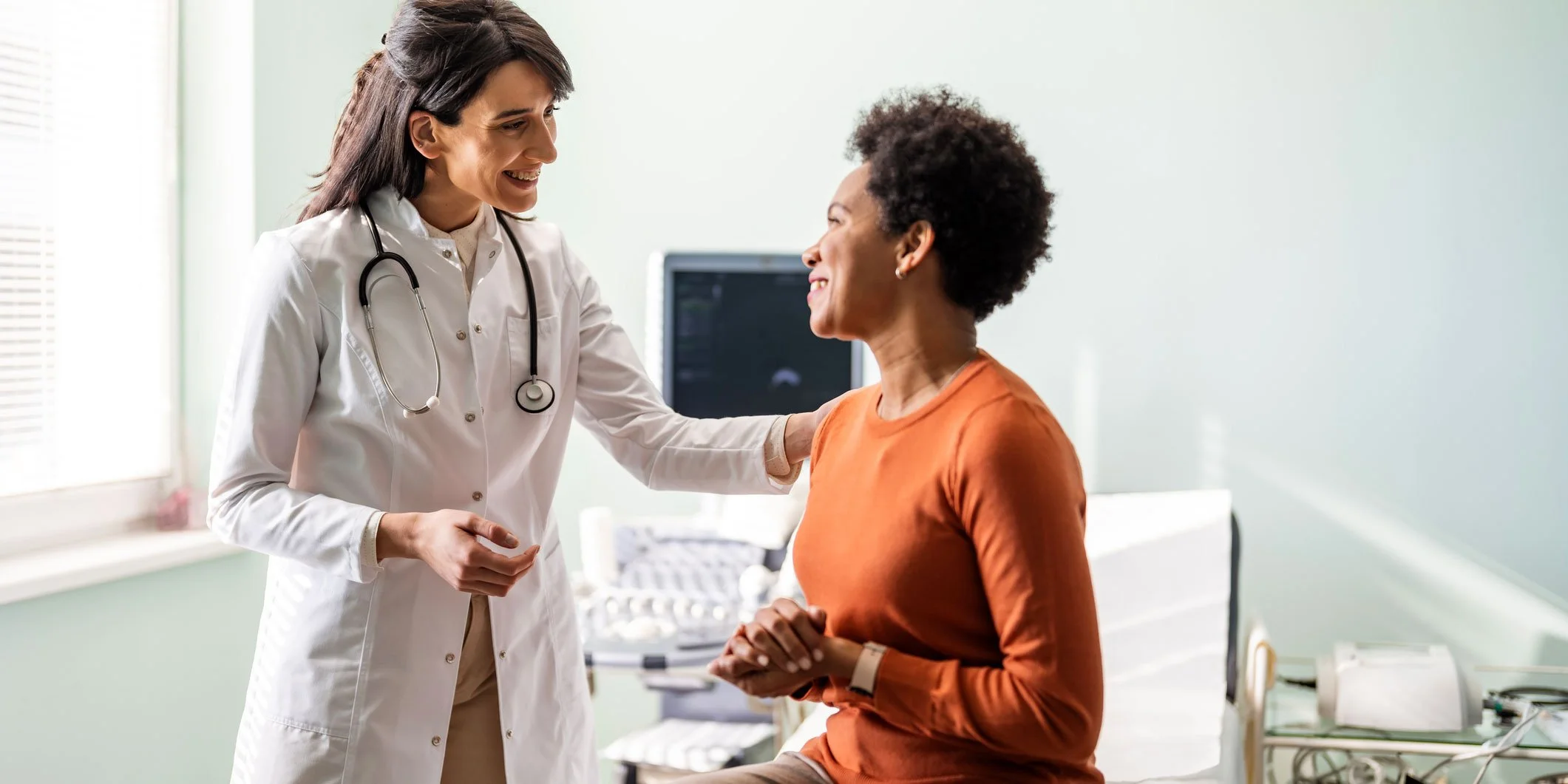 A female doctor and a female patient smiling and talking in a medical examination room.