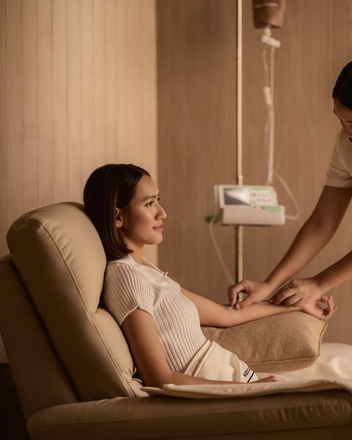 A woman receiving an Energise and Revitalise IV drip in a clinical room, sitting on a beige recliner chair, with a healthcare professional adjusting her IV.