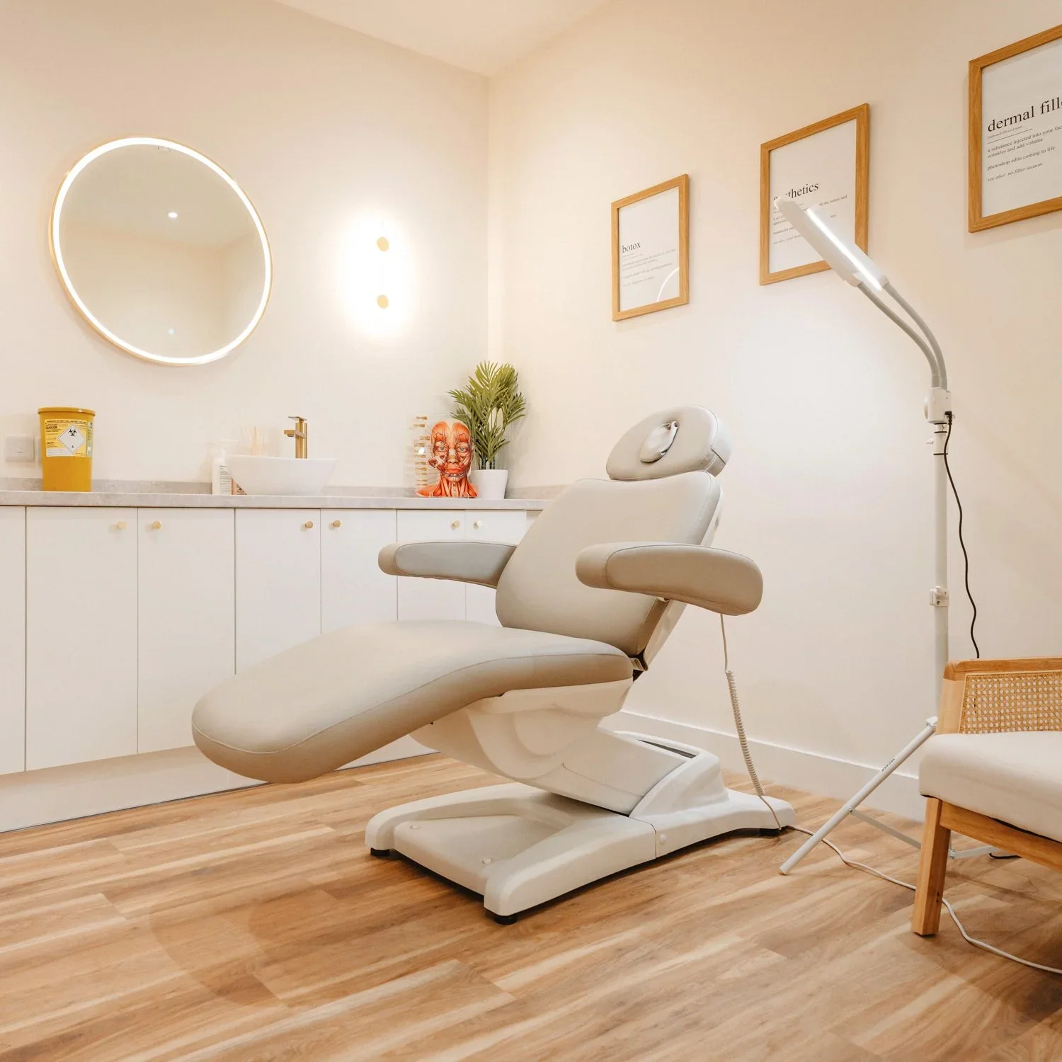 Medical treatment room with a white examination chair, a wall mirror, framed informational posters, and a countertop with a sink, plant, and decorative items.