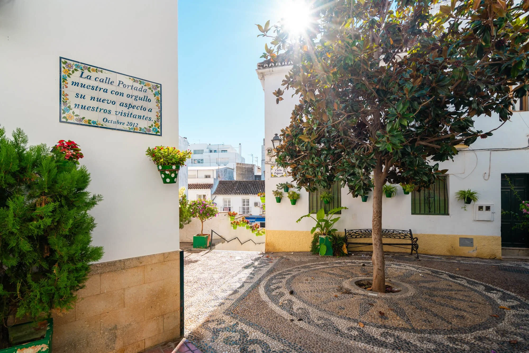 A white-walled outdoor area with a decorated street sign, potted plants, and a large leafy tree. The sign reads 'La calle Portada muestra con orgullo su nuevo aspecto a nuestros visitantes. Octubre 2012.' The sun shines brightly overhead, casting light on the scene.