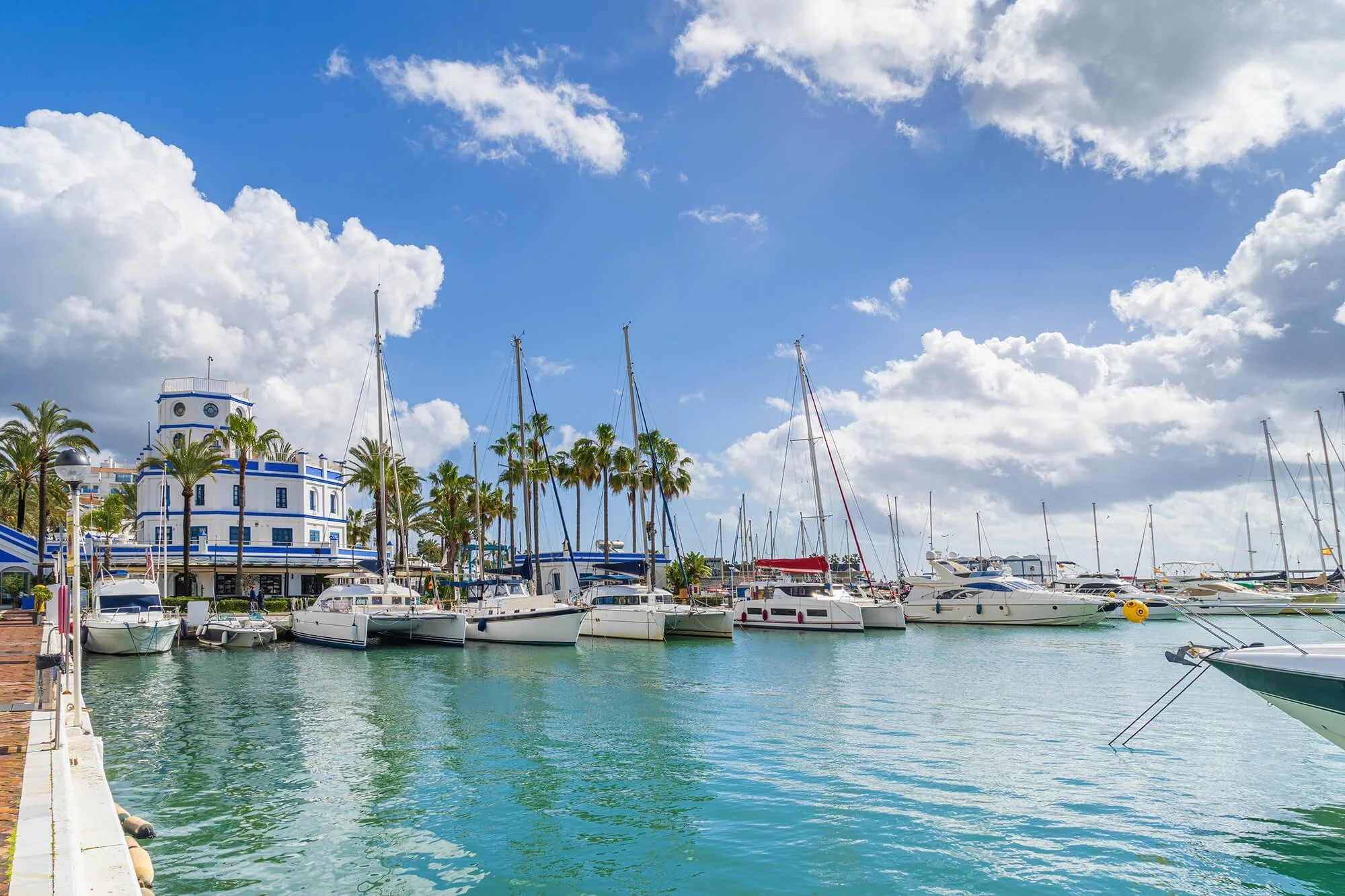 Marina with sailboats docked along the pier, palm trees, a blue and white building, and a partly cloudy sky.