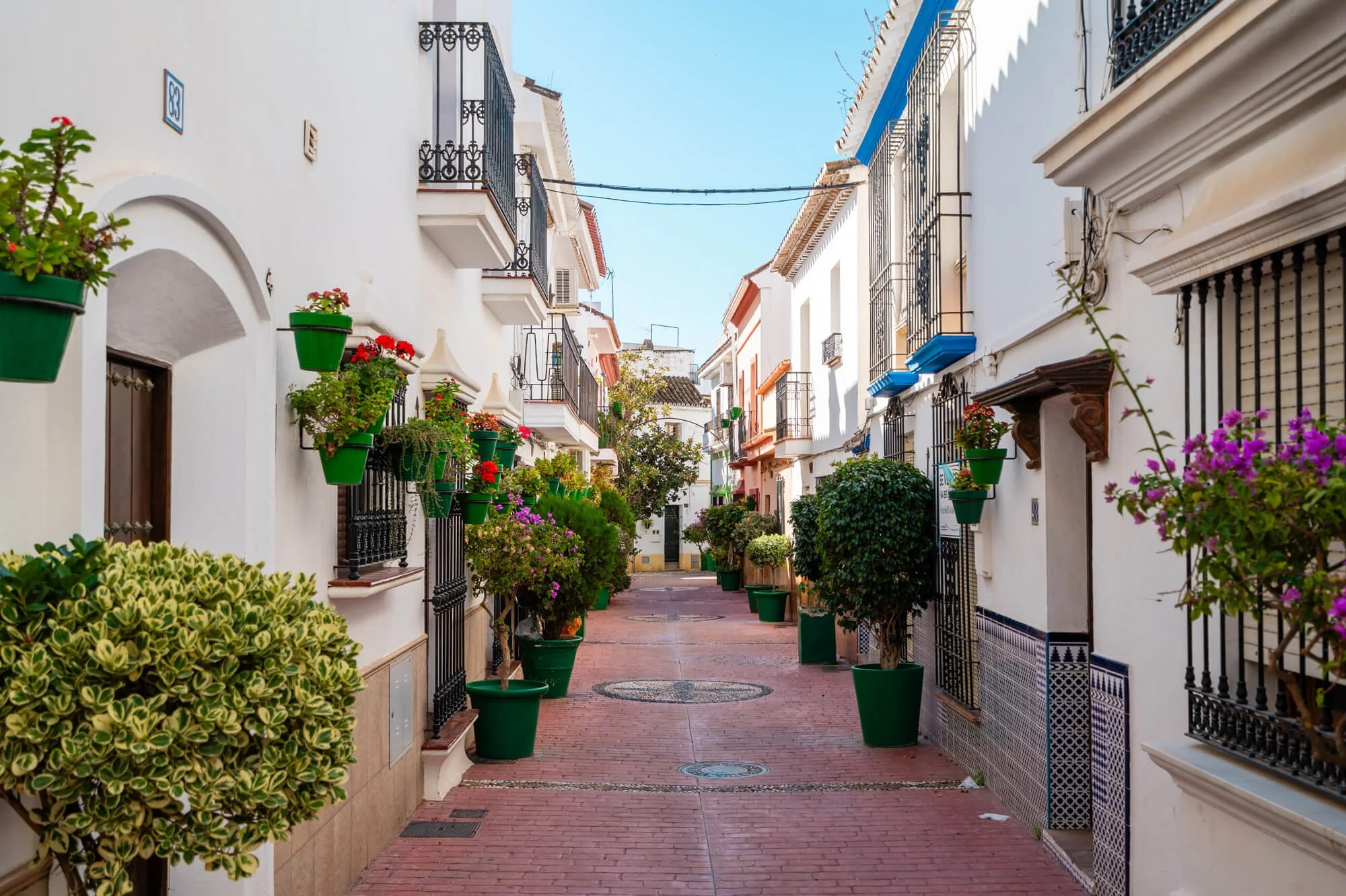 A narrow street in a Mediterranean town lined with white buildings decorated with colorful flowers in green pots. The street is paved with red bricks, and the buildings have balconies with black wrought iron railings and some with blue accents. Clear blue sky and sunny weather are visible.