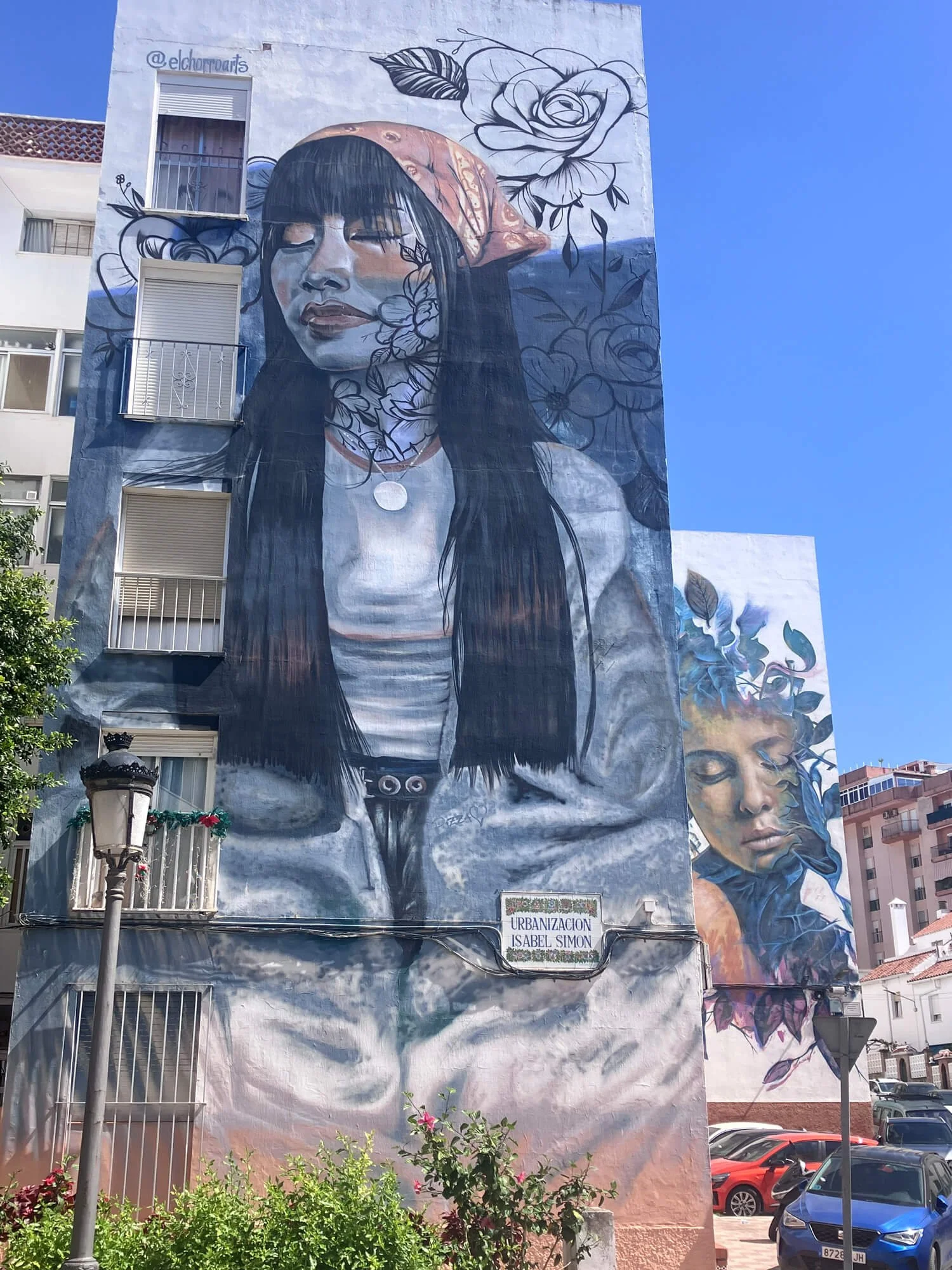 Large mural on building featuring a portrait of a woman with long dark hair, surrounded by floral designs and a large rose. The mural includes the text '@elchoruants' and a sign that reads 'Urbanizacion Isabel Simon' beneath it.