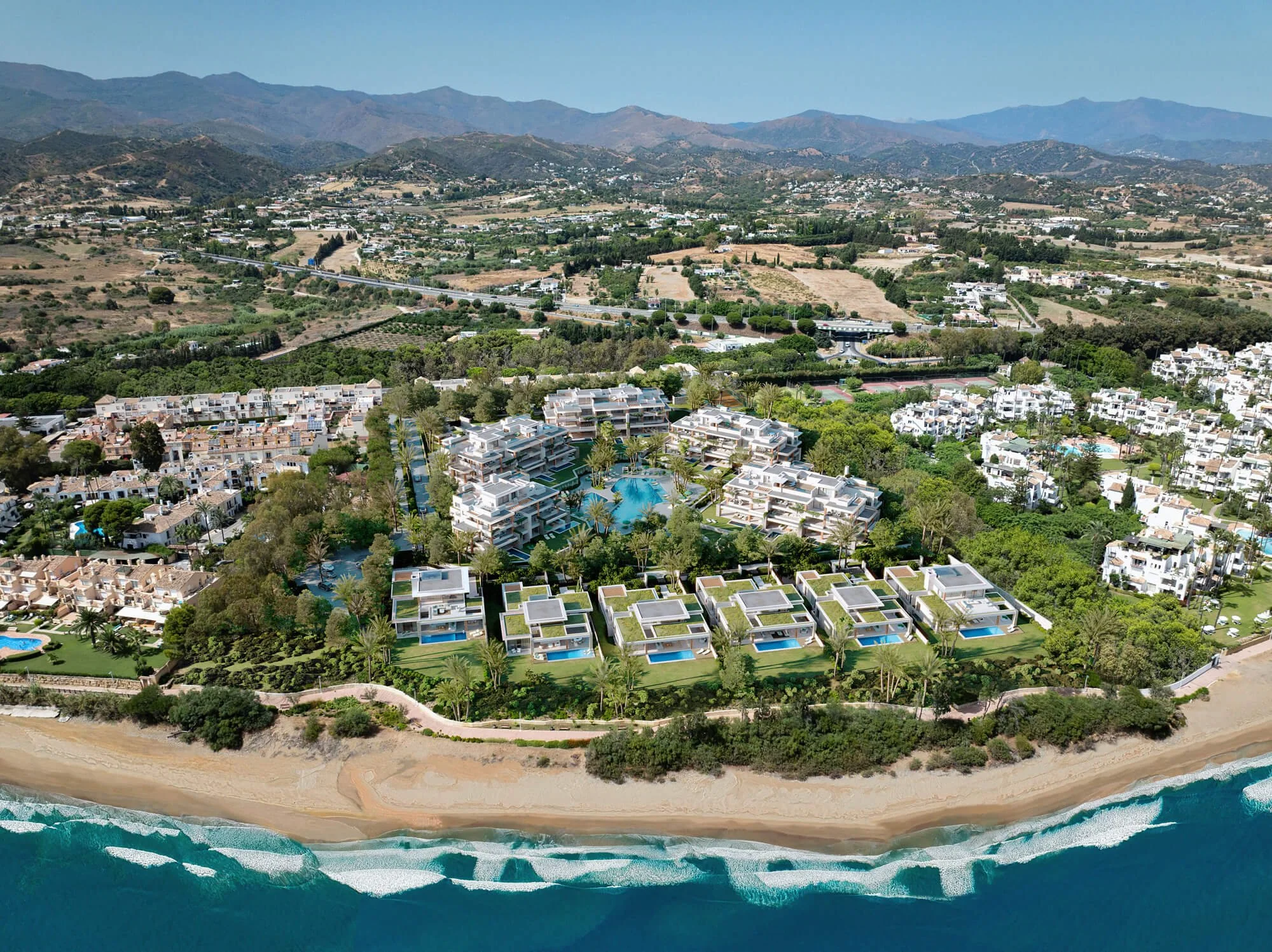 An aerial view of a coastal resort with white buildings, swimming pools, lush greenery, and the ocean with waves crashing on the sandy beach.