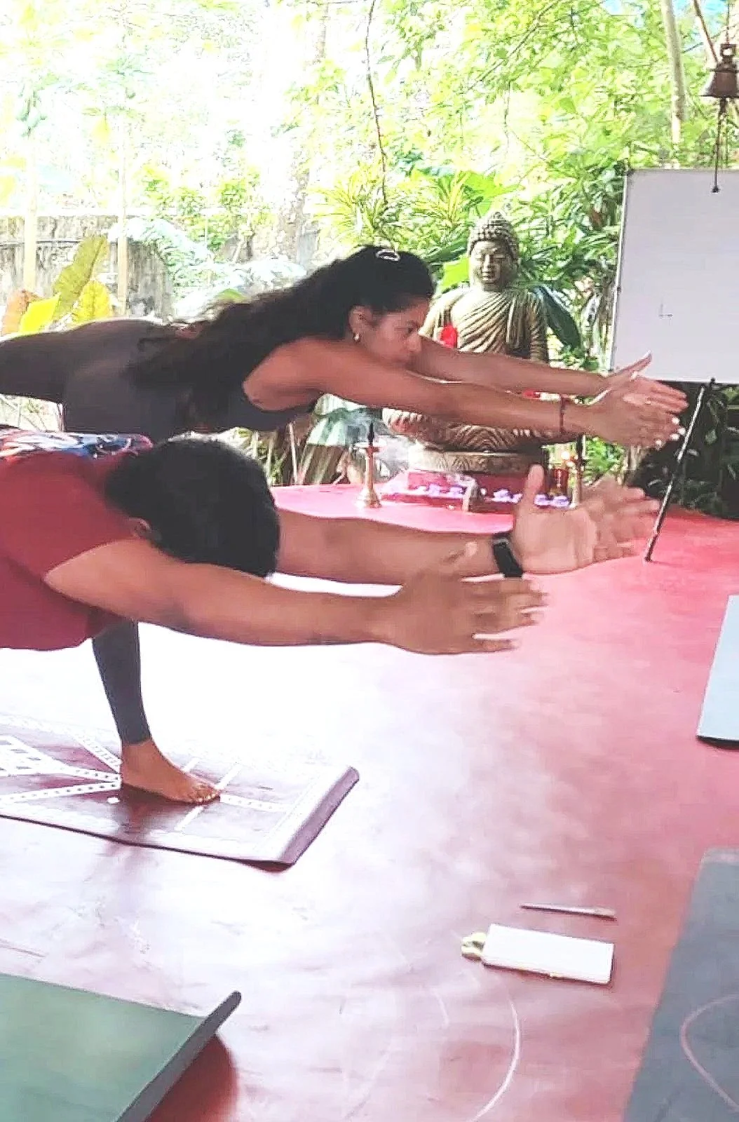 Two people practicing yoga outdoors, both in forward bent positions with arms extended. One is on a yoga mat, and the other is on a yoga block. There are statues and greenery in the background.