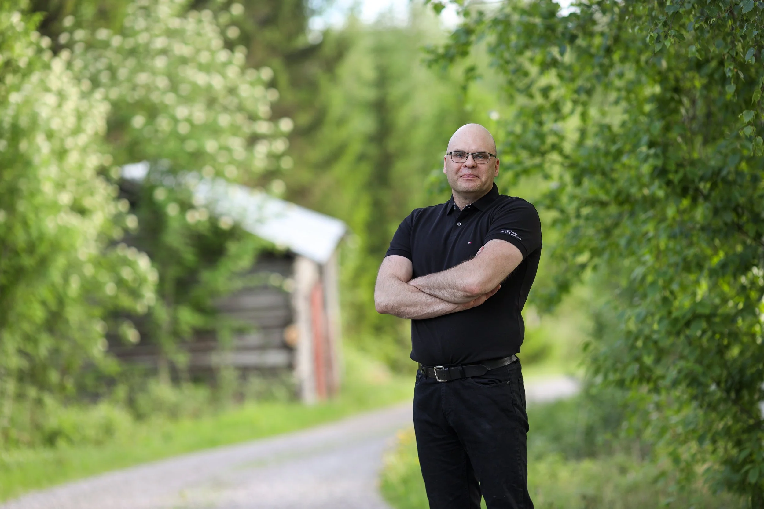 Timo Pajala seisoo soratiellä mustassa pikeepaidassa ja mustissa housuissa. Taustalla koivuja ja vanha lato.