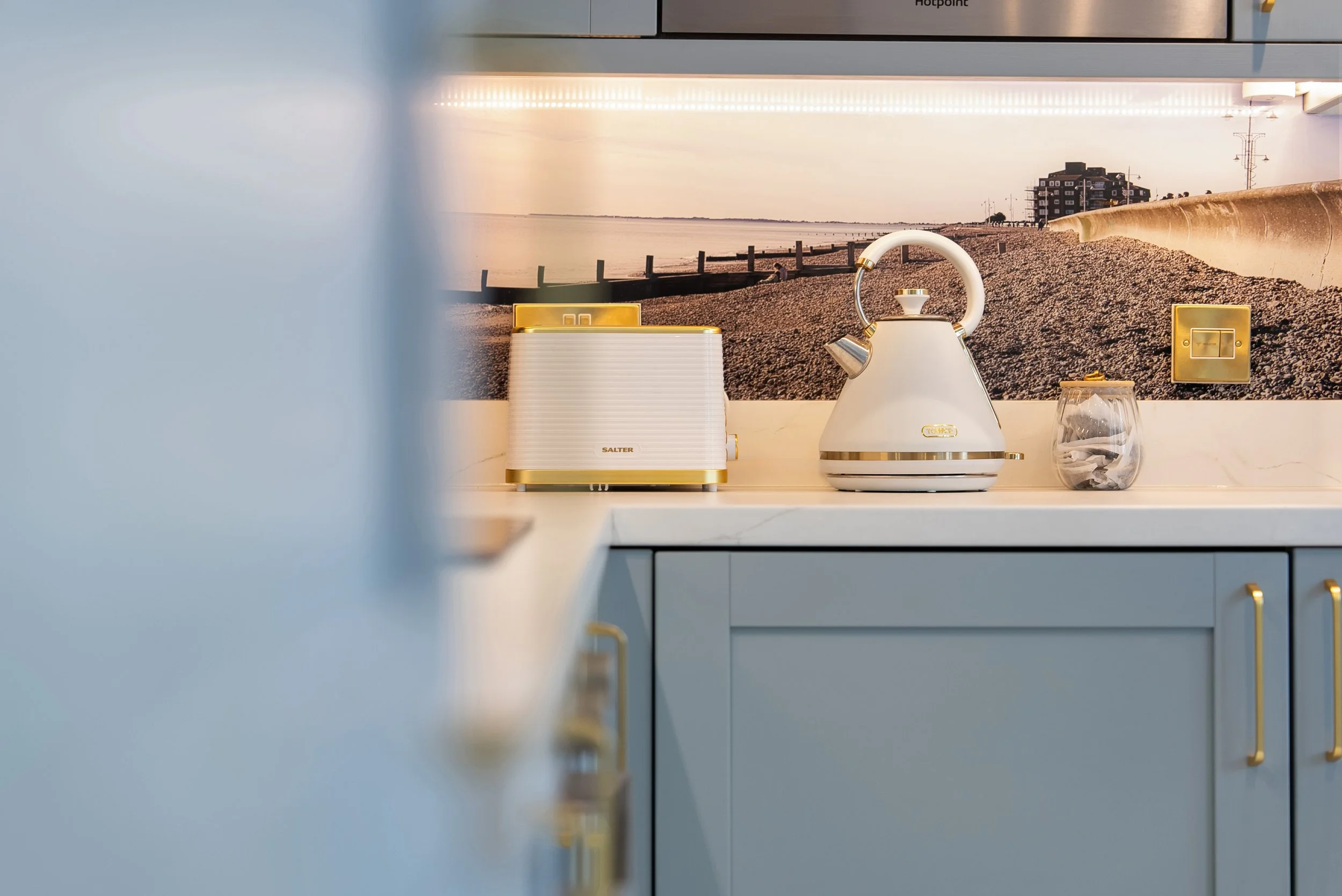 Kitchen counter with a white and gold kettle, a white toaster, a glass container filled with tea bags, and a wall decal of a beach scene with buildings and power lines.