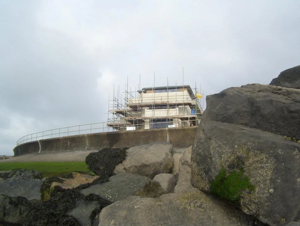 Building under construction on a hill, surrounded by scaffolding and rocks, with cloudy sky in the background.