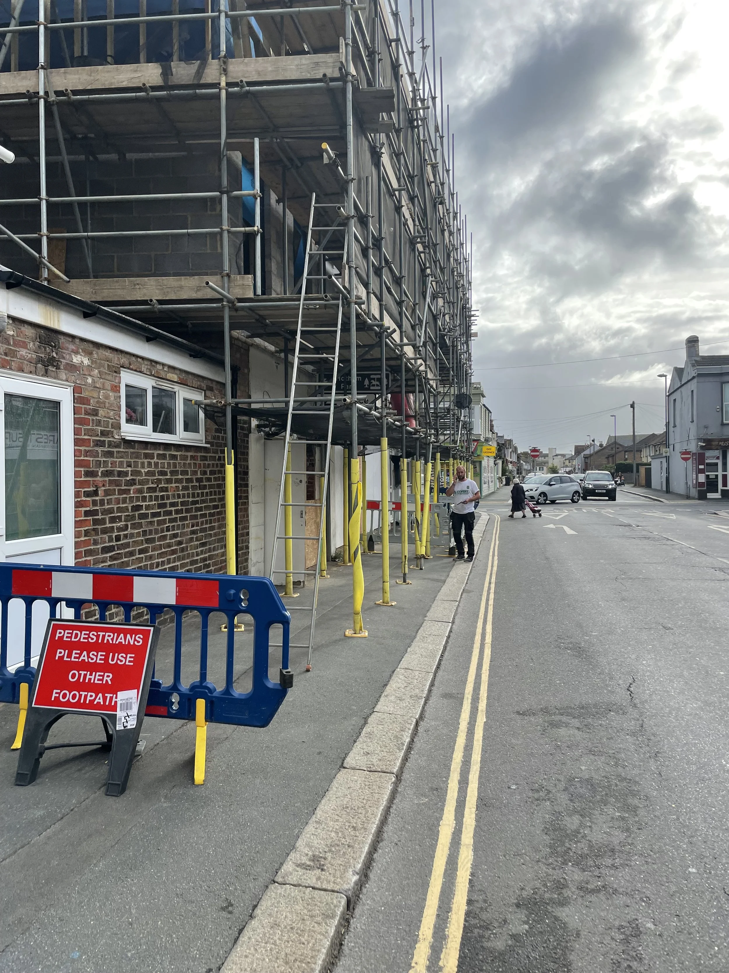 Construction scaffolding around a multi-story brick building under renovation with a street, pedestrians, and parked cars nearby.