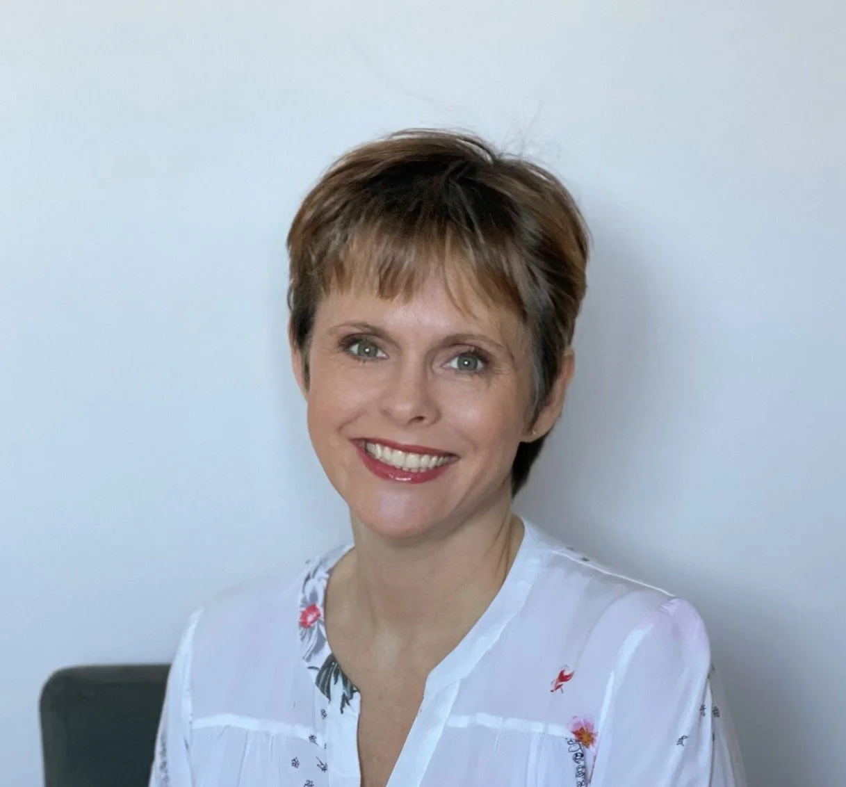 A woman with short brown hair and blue eyes, smiling, wearing a white blouse with embroidered floral patterns, sitting against a plain light-colored wall.