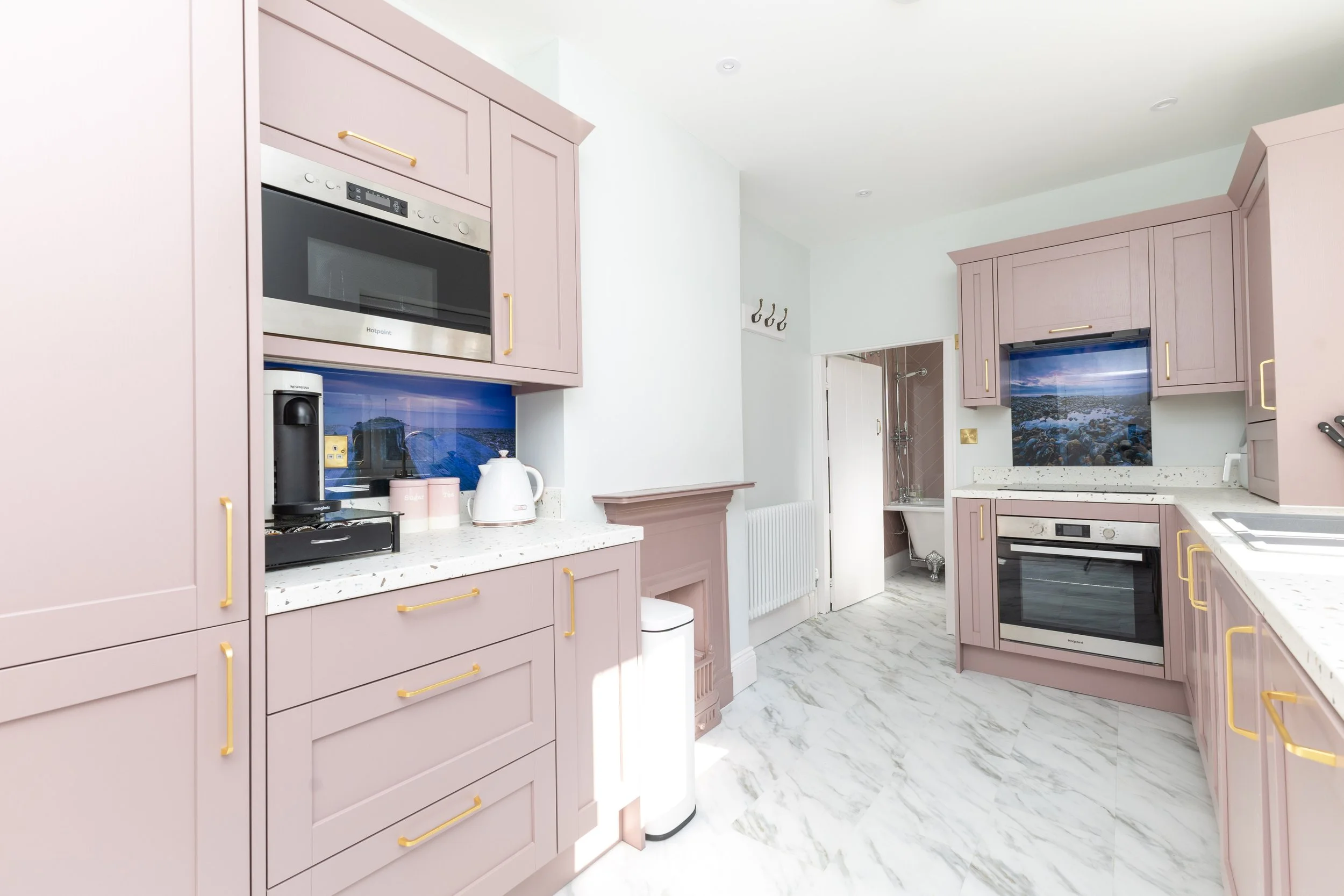 Kitchen with pink cabinets, white countertops, and a marble floor. There is a built-in microwave, a coffee maker, a kettle, and a stove. The kitchen has a backyard view through the windows.