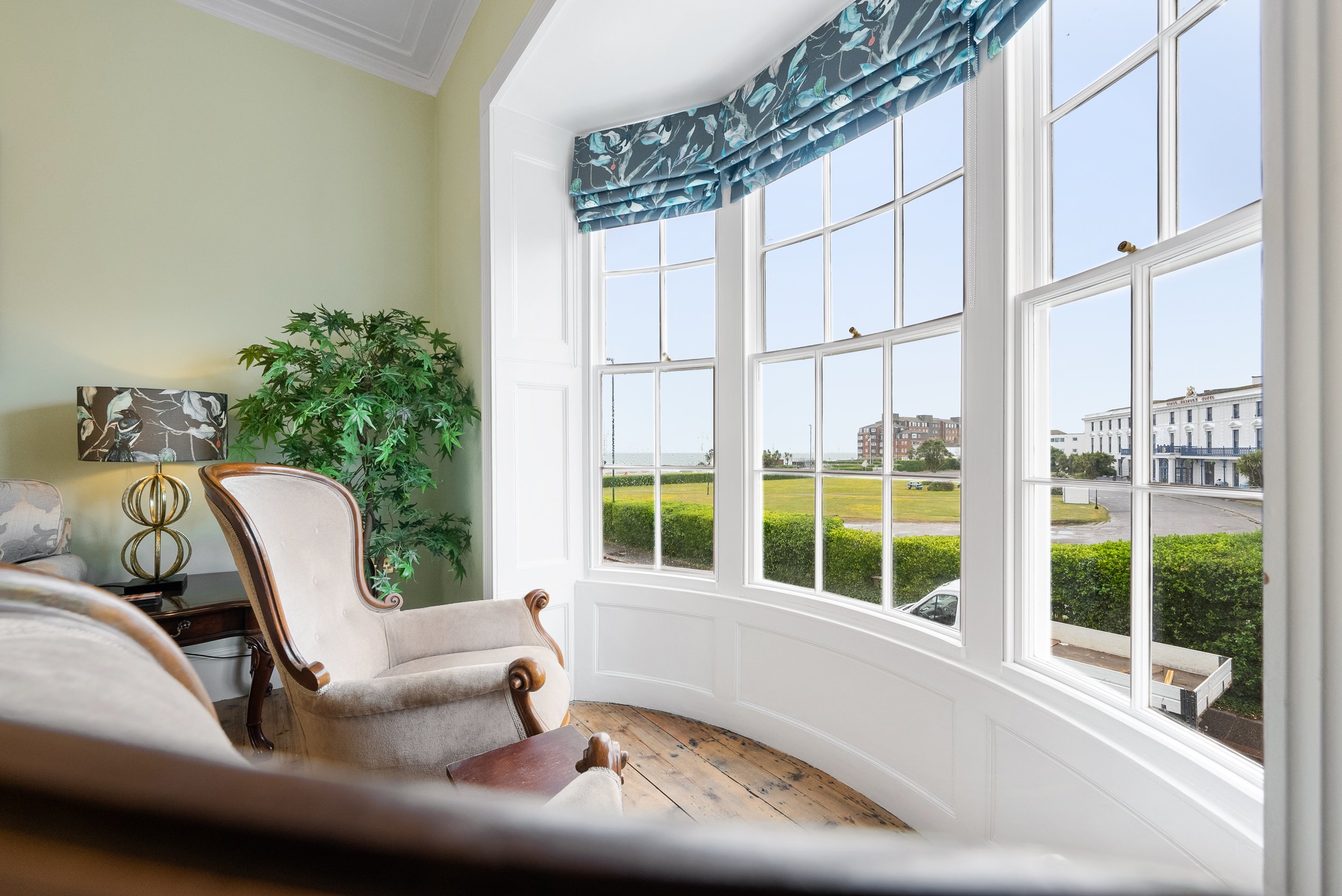 Interior of a room with beige furniture, a lamp with a black and floral shade, green houseplant, and large bay window showing a view of a suburban street and blue sky.