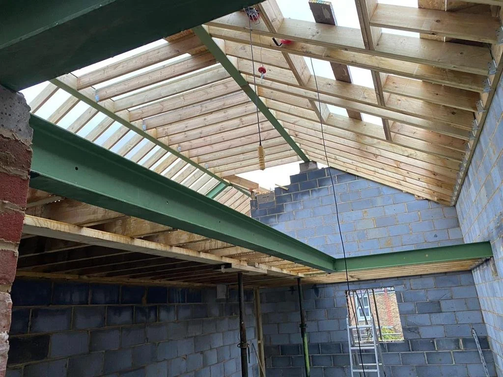 Construction site inside a building with exposed wooden roof framing, steel beams, brick walls, and a ladder.