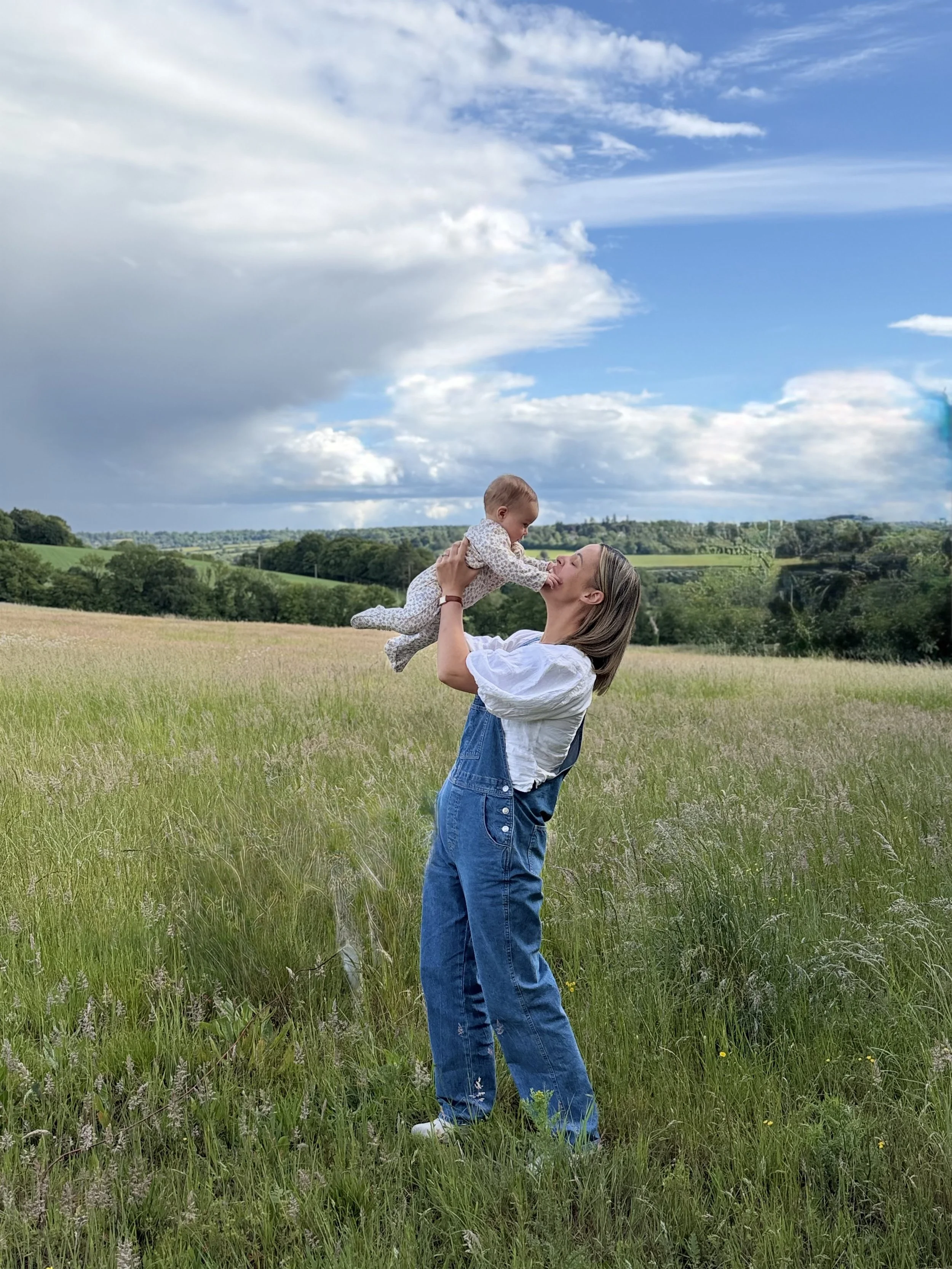 A woman holding a baby girl in a grassy field, with a landscape of trees and hills and a partly cloudy sky in the background. Thoughtful, skincare with purpose.