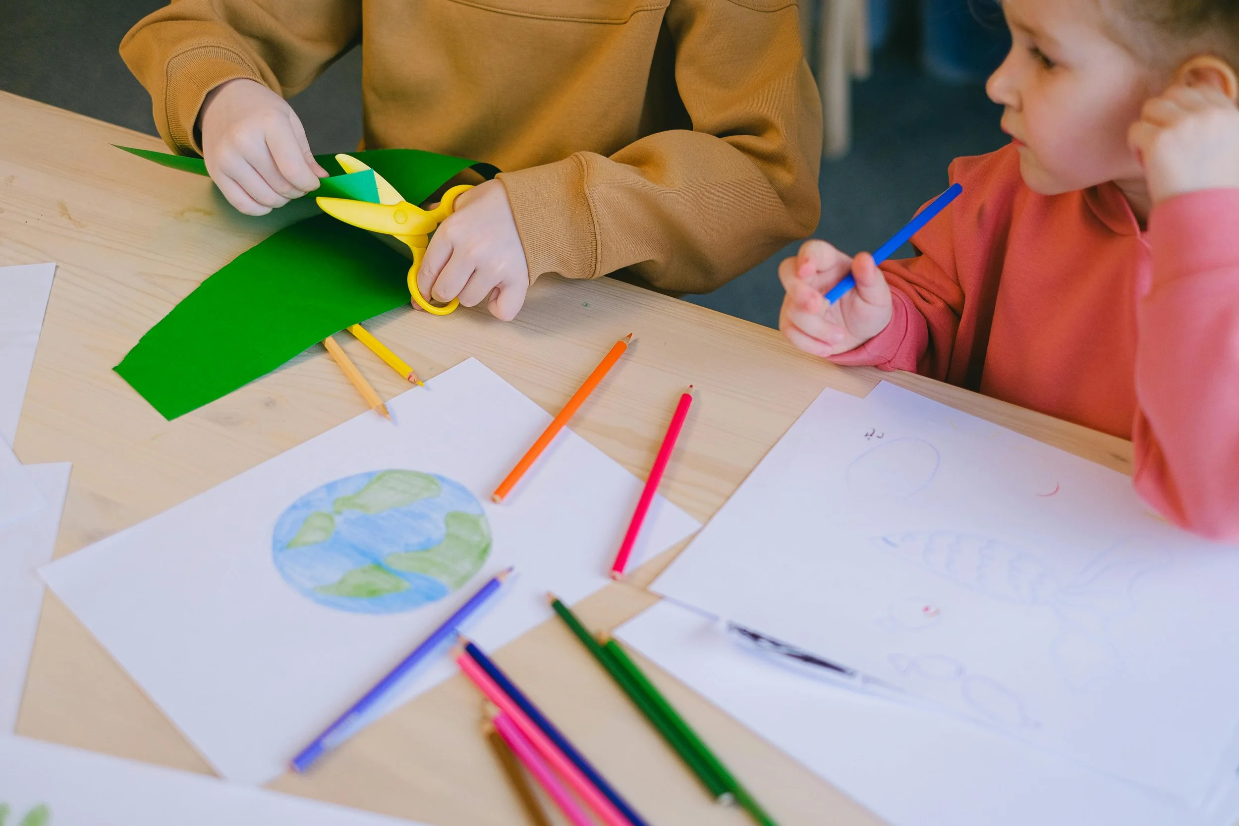 Two children sitting at a table, coloring and cutting paper crafts. One child is using scissors with yellow handles to cut green paper, and the other is holding a blue marker, with drawings and colored papers on the table.
