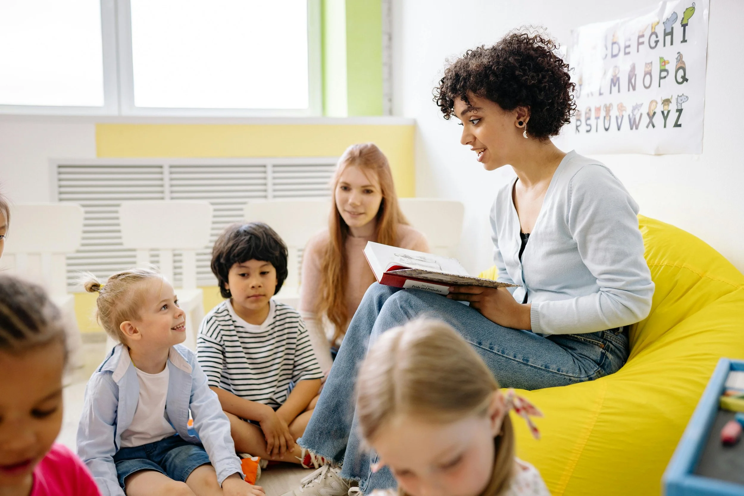 A classroom scene showing a teacher reading to young students who are seated on the floor, with a colorful alphabet poster on the wall and sunlight coming through the windows.