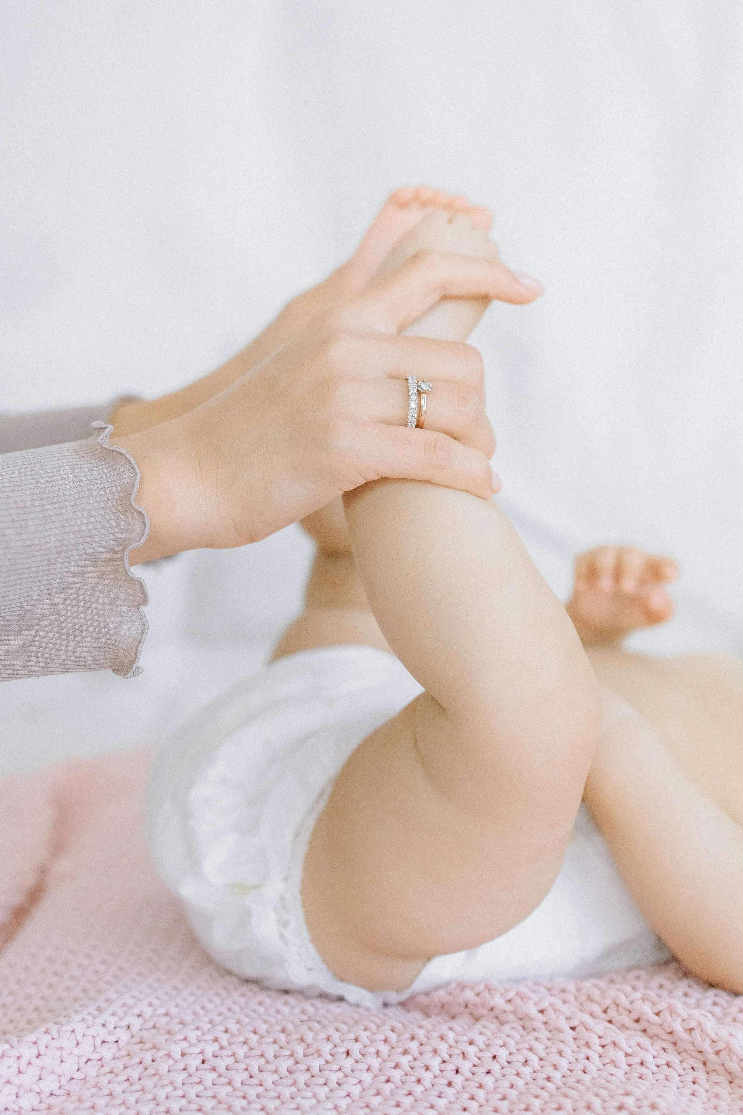 A baby lying on a pink knitted blanket, with an adult's hand gently holding the baby's leg. The hand is wearing a ring with diamonds, and the baby is wearing a white diaper.