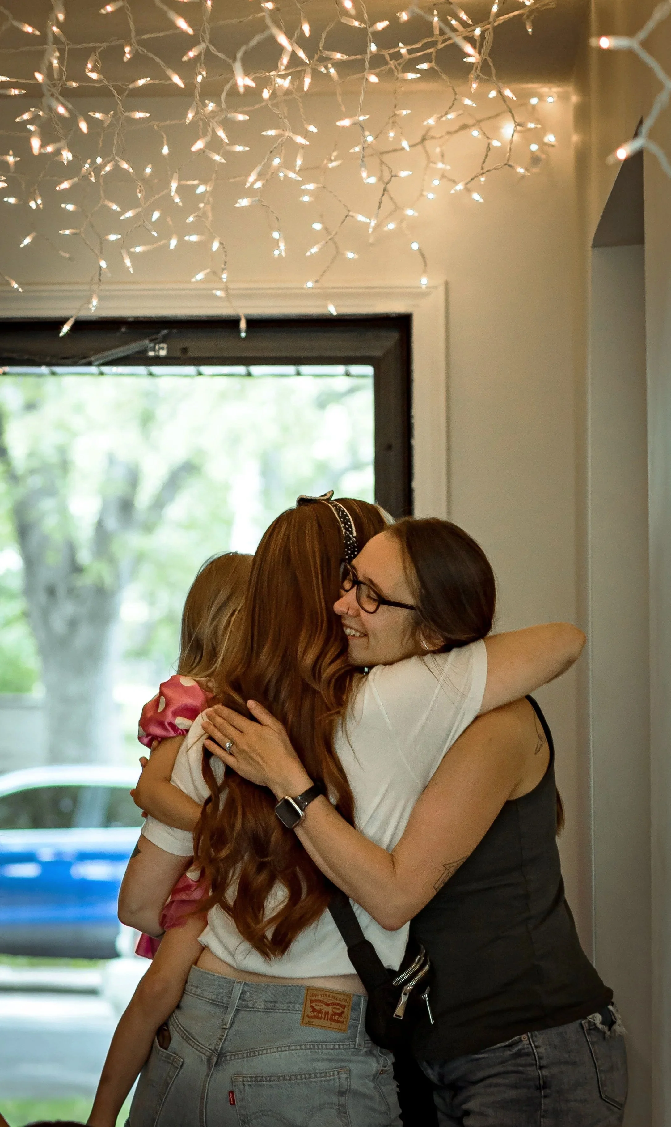 Two women hugging each other inside a room, with string lights hanging from the ceiling. The background shows a window with trees outside.