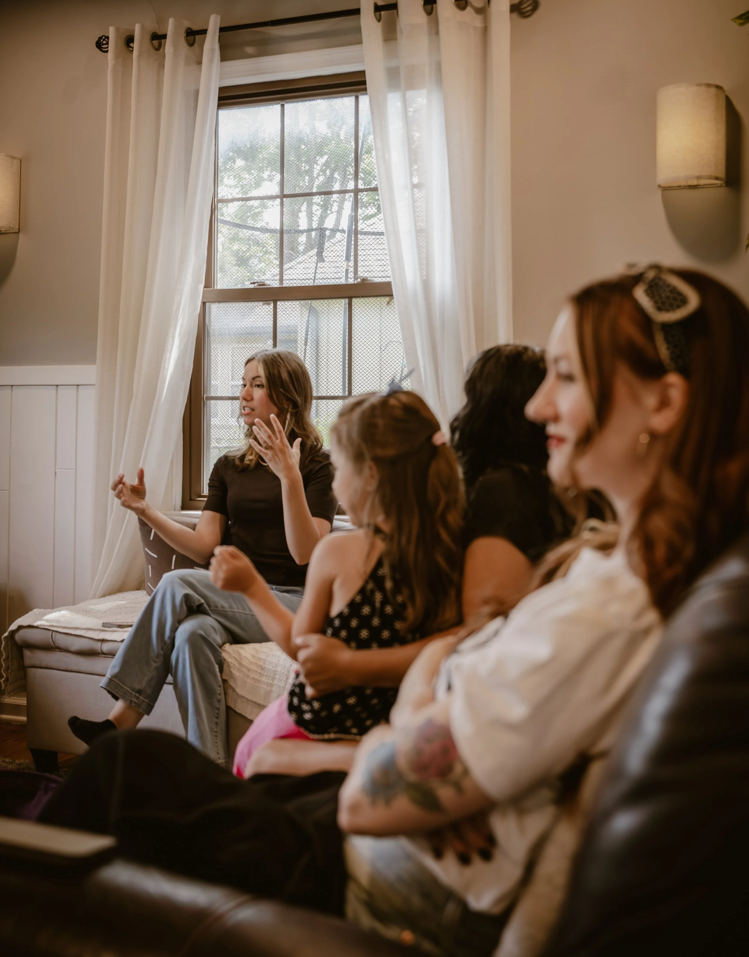 A group of people sitting in a cozy room with large window, white curtains, and beige walls, engaging in conversation.