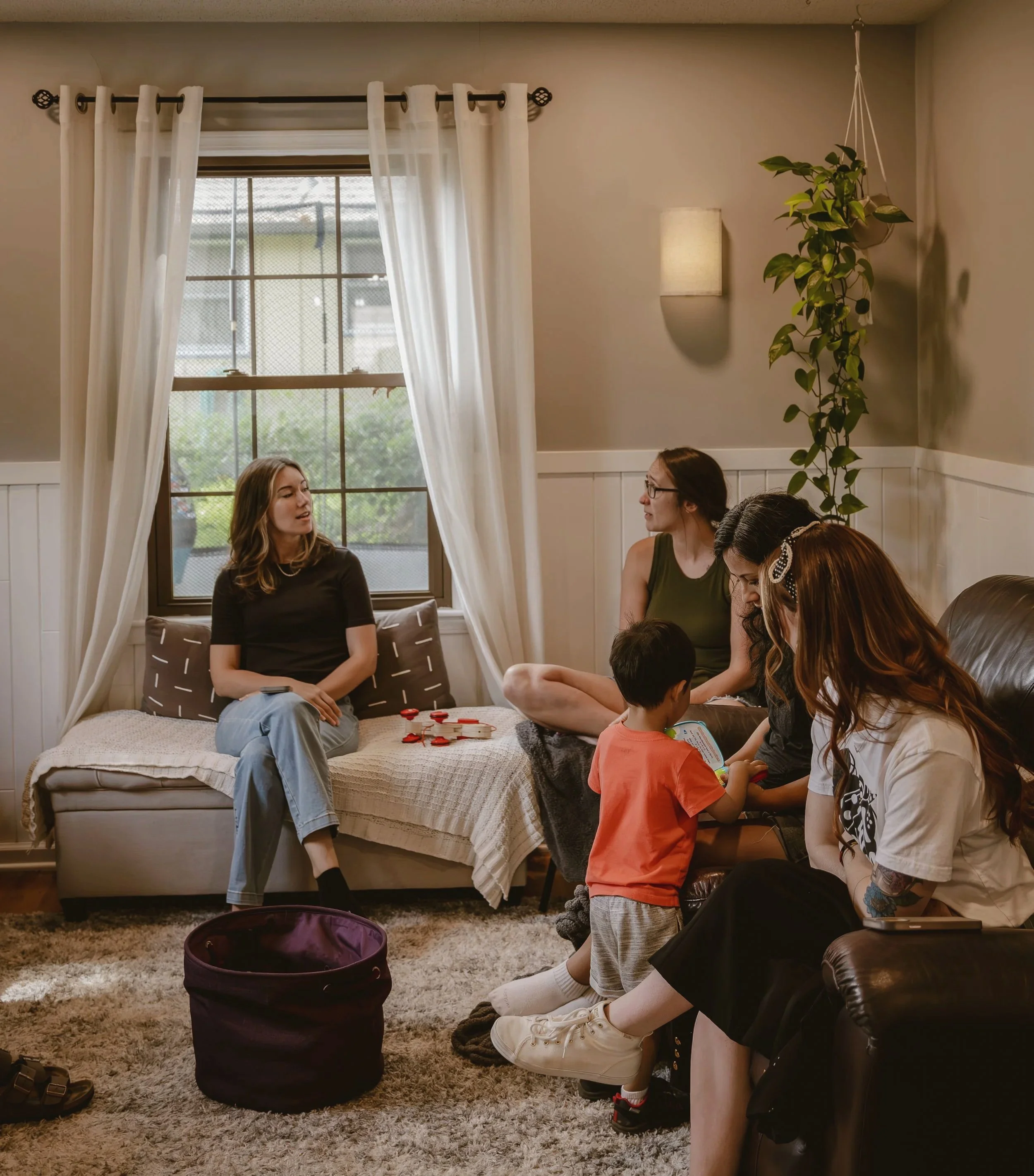 Group of people sitting together in a cozy living room, engaged in conversation. There's a large window with white curtains, a hanging plant, a wall lamp, and a shaggy carpet on the floor.