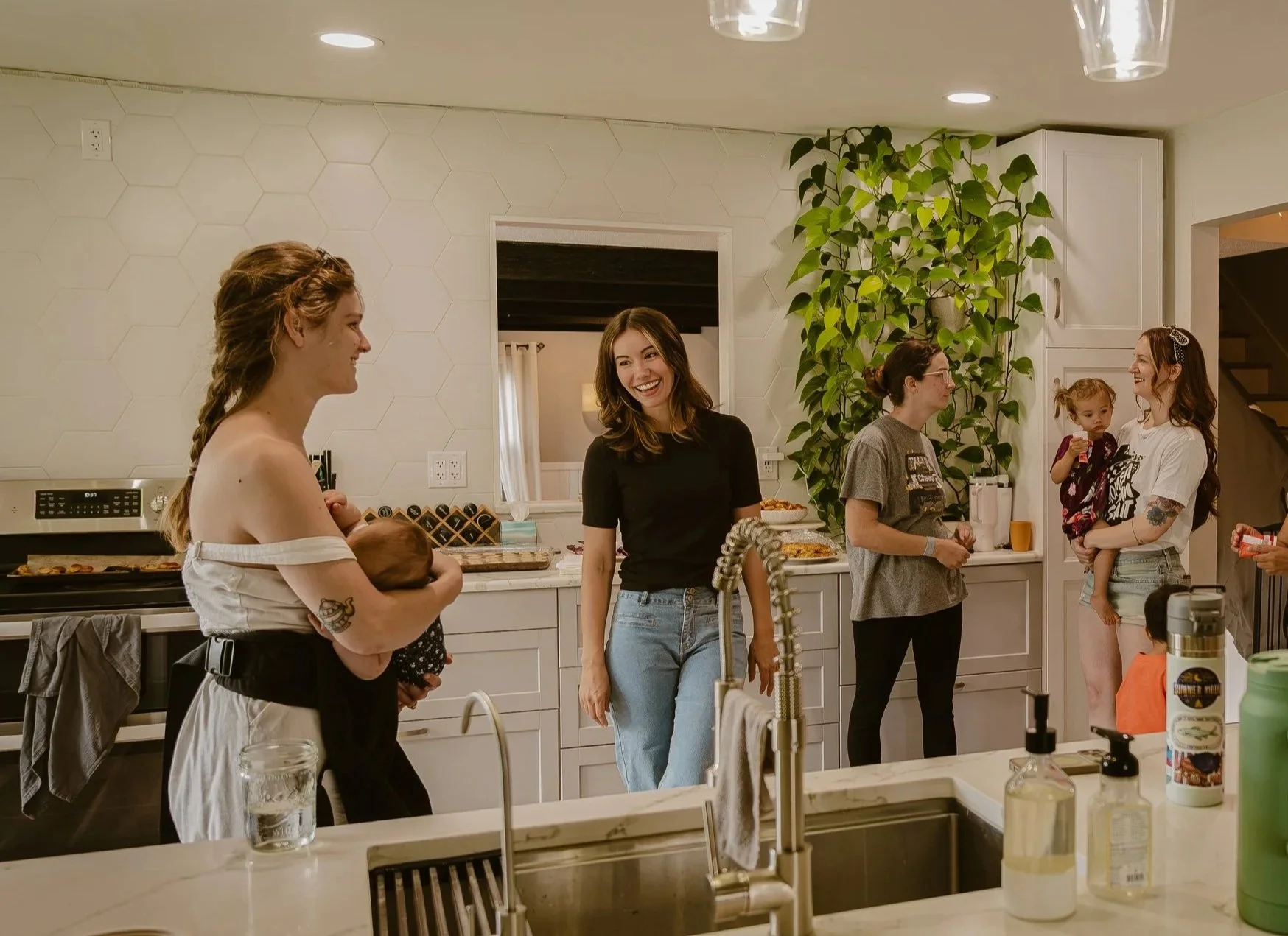 Group of women and a young girl gather in a modern kitchen, socializing and smiling, with a large green plant in the background, and a countertop with various items including drinks and snacks.