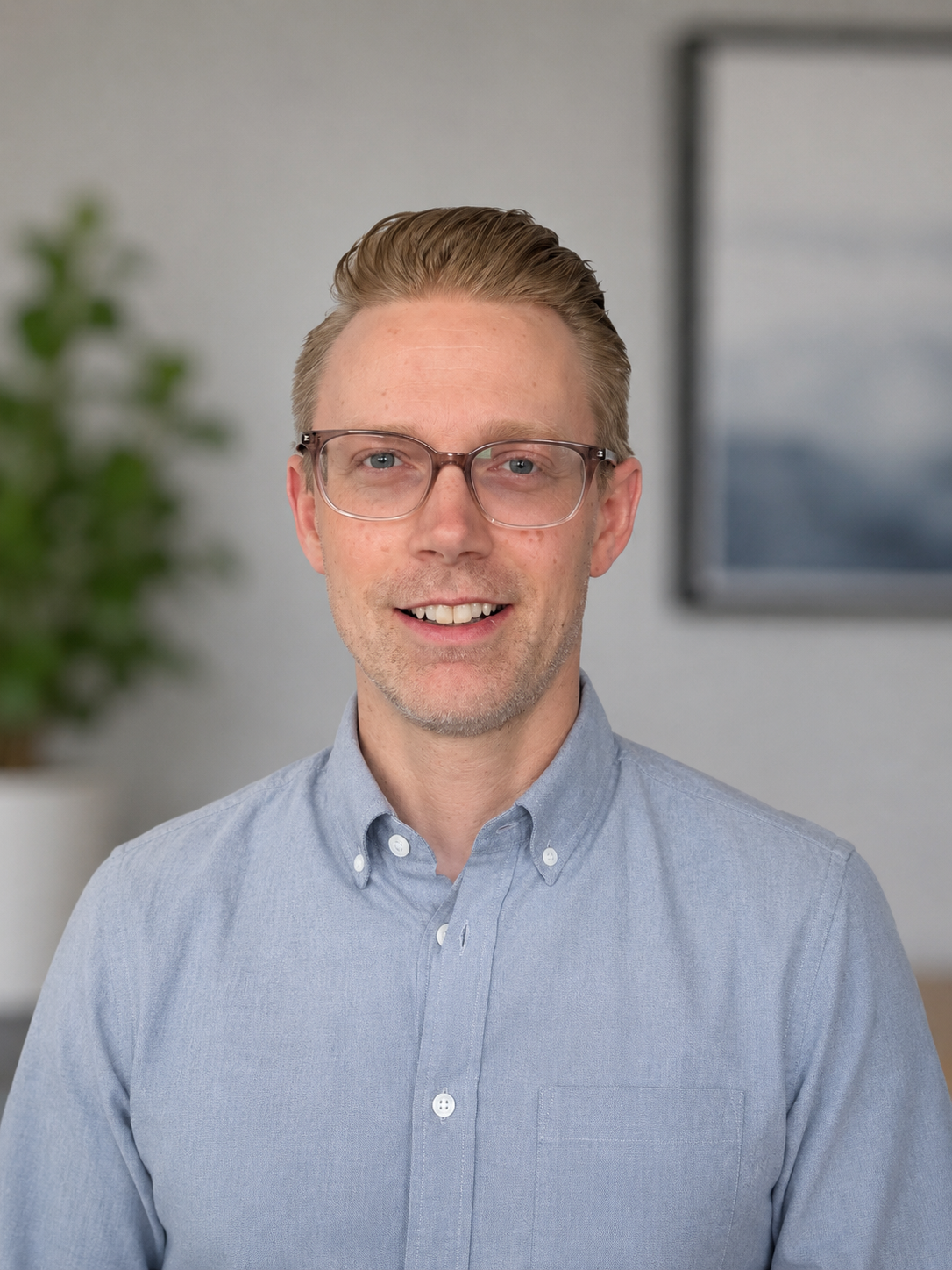 A man with light brown hair, glasses, and a light blue button-up shirt, smiling in an office setting with a blurred plant and framed picture in the background.