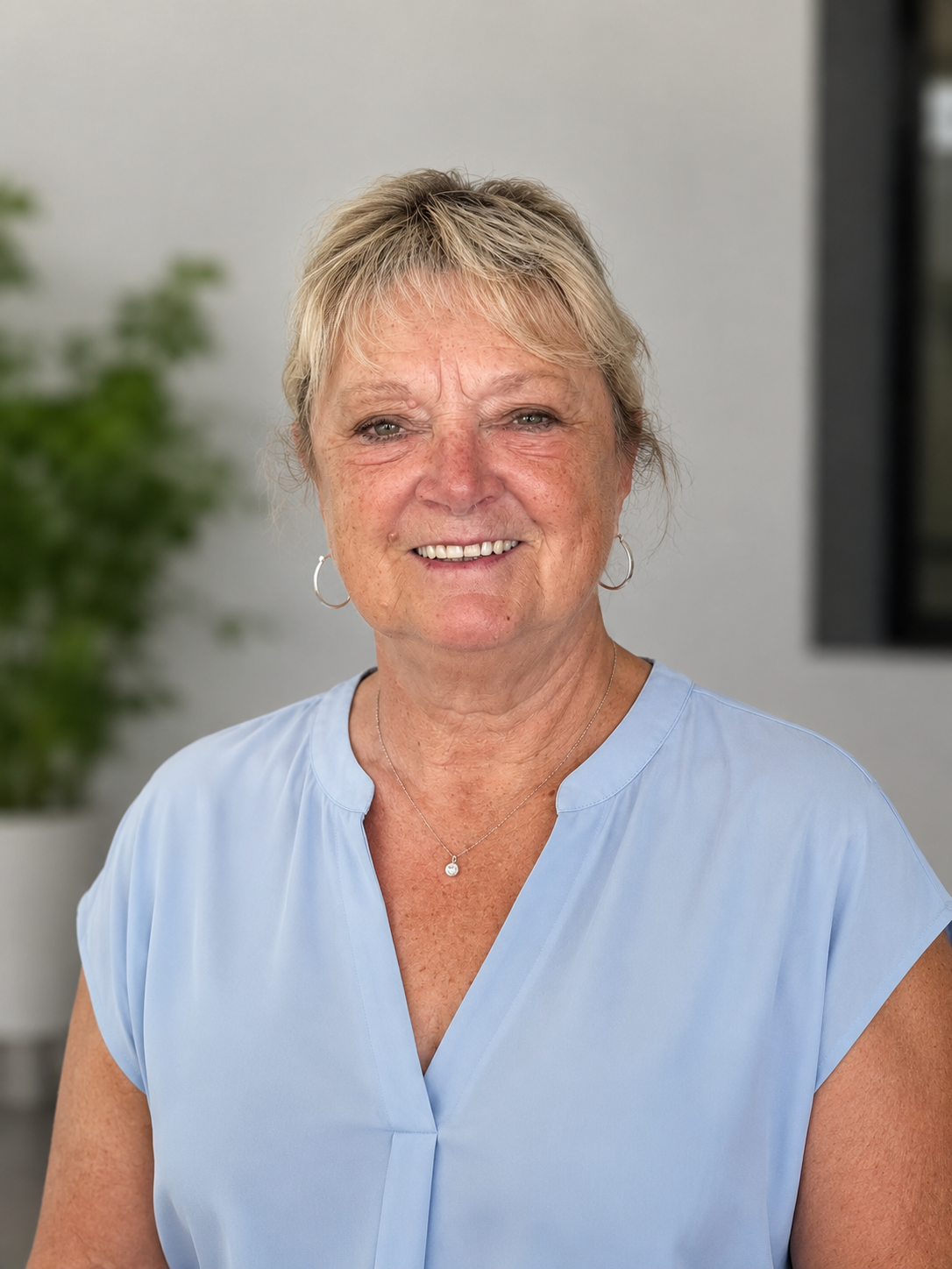 Portrait of a middle-aged woman with short blonde hair, wearing a light blue blouse, pearl necklace, and hoop earrings, smiling in front of a blurred background.
