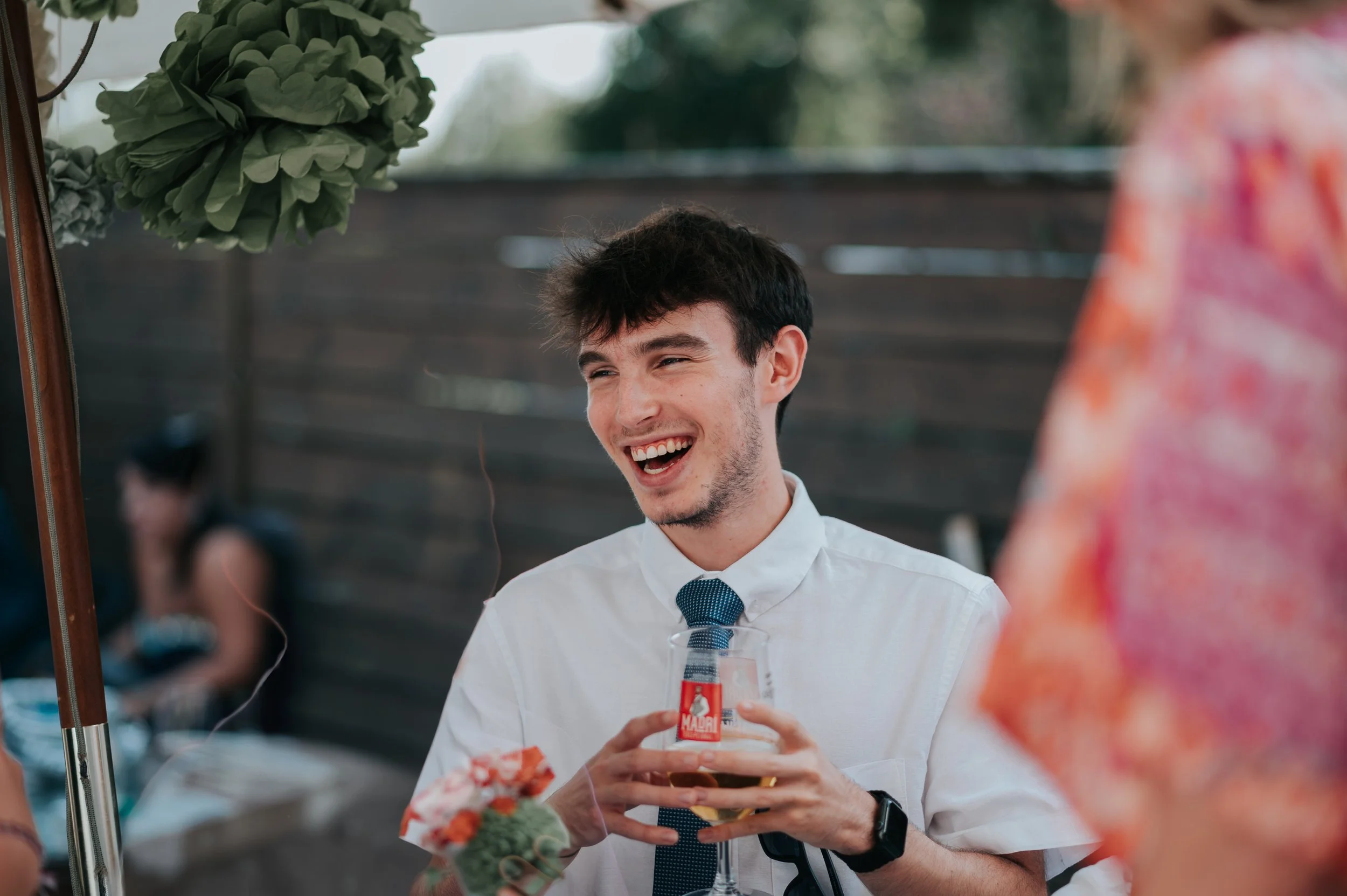 A young man wearing a white dress shirt and a blue tie is smiling and holding a glass of beer during a social event or gathering.