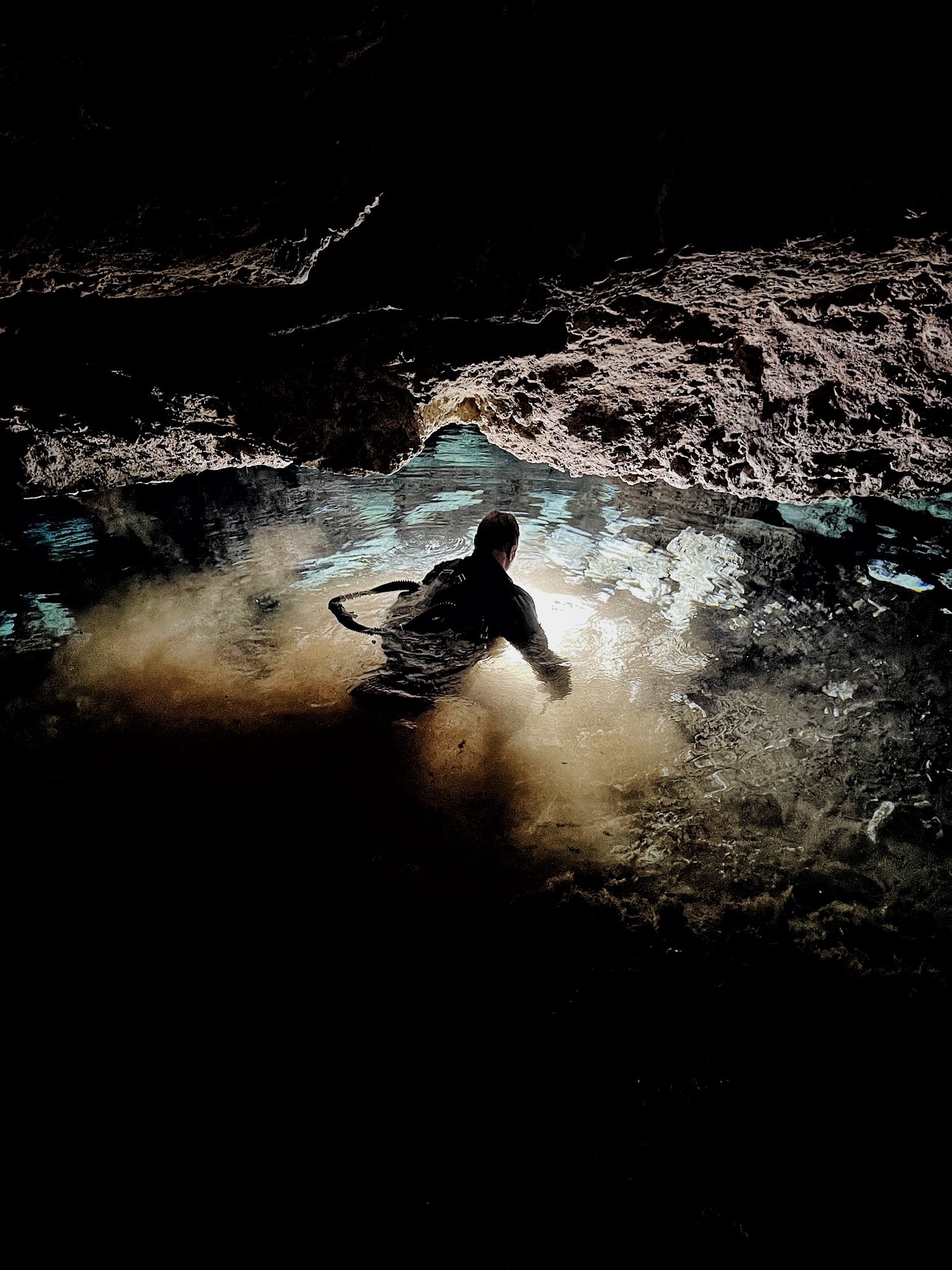 Person exploring a cave with a small pool of water, rocky walls, and an opening in the distance letting in light.