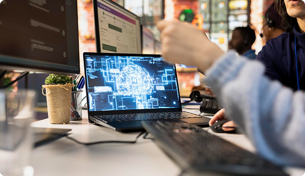 Close-up of a laptop displaying a digital brain and various data graphics, with a person pointing at the screen, in a collaborative workspace.