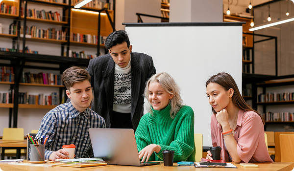 Four young people gathered around a laptop at a table in a library or bookstore, with bookshelves in the background. They are working together, smiling, and looking at the laptop screen.