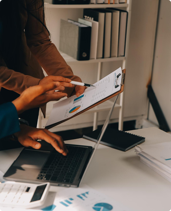 Two people reviewing documents with charts and graphs in an office, one using a laptop and the other pointing at the paper.