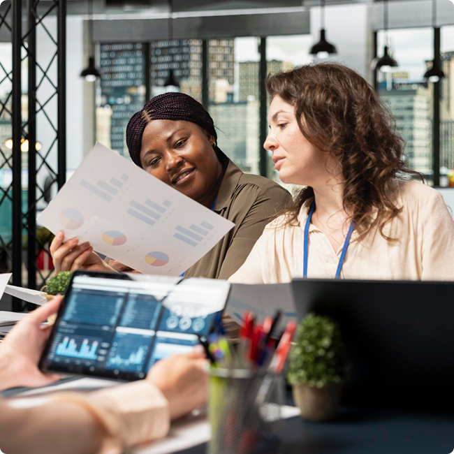 Two women in an office discussing data charts, with a person in the foreground working on a laptop with data graphs, in a modern high-rise office with city views.