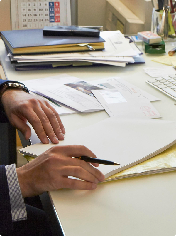 A person with a business suit holding a pen and writing or signing a document at a cluttered office desk with papers, folders, and office supplies.