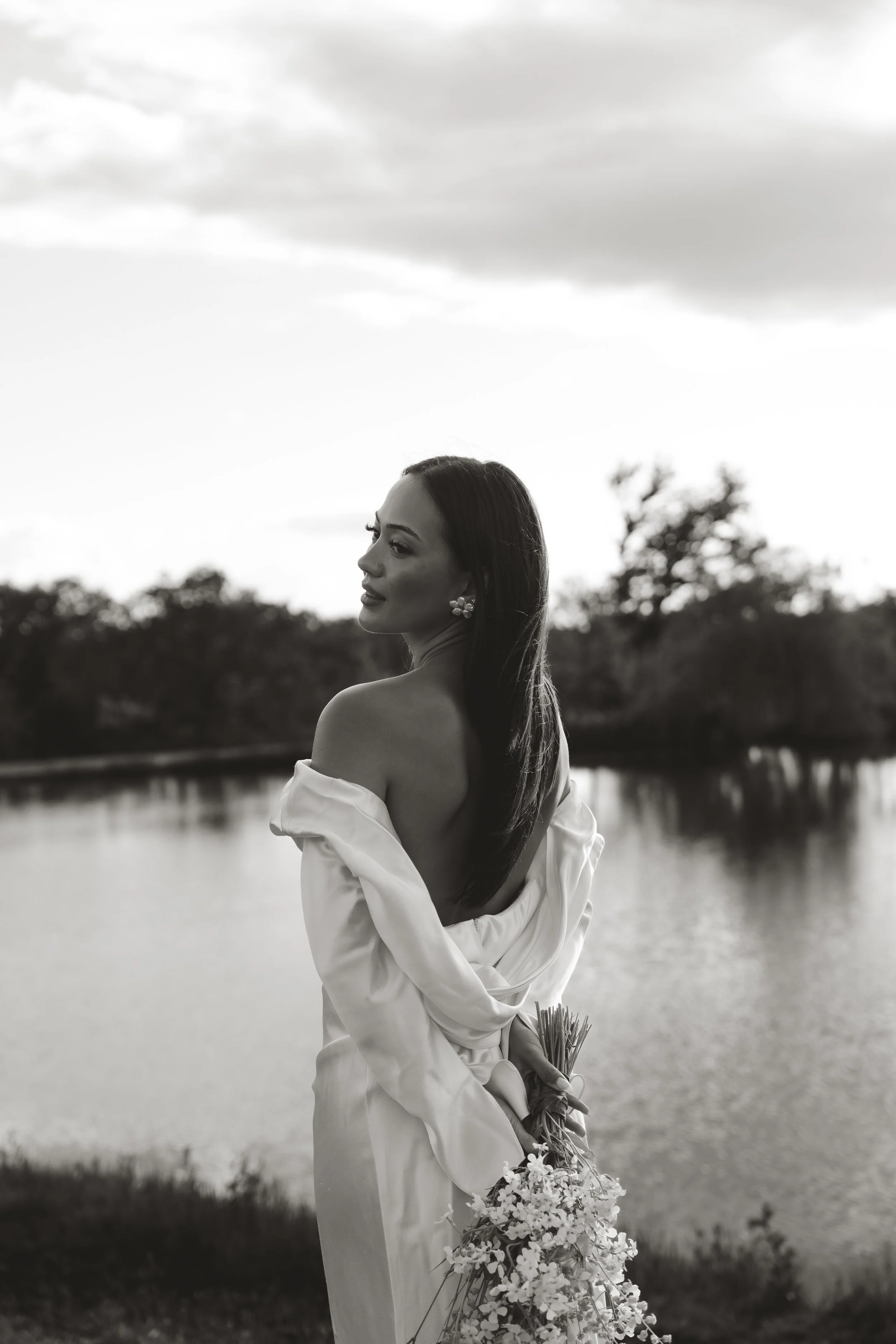 An elegant bride standing outdoors near a body of water holding a bouquet of flowers, with trees and a cloudy sky in the background, in black and white.