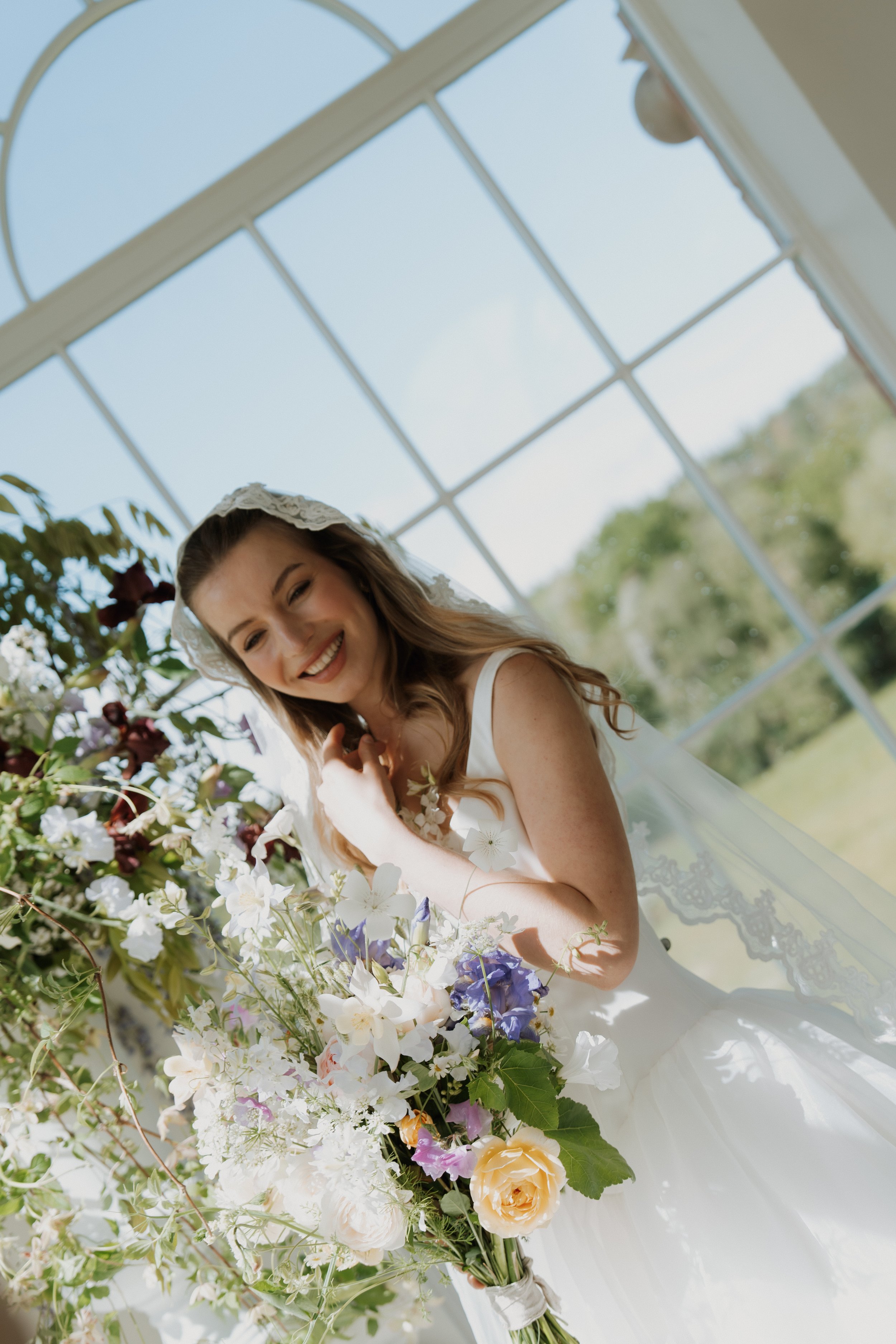 A smiling bride with long, wavy hair wearing a luxury Savannah Miller Bridal wedding gown and lace veil, holding a large bouquet of white, purple, and yellow flowers by a sunlit window.