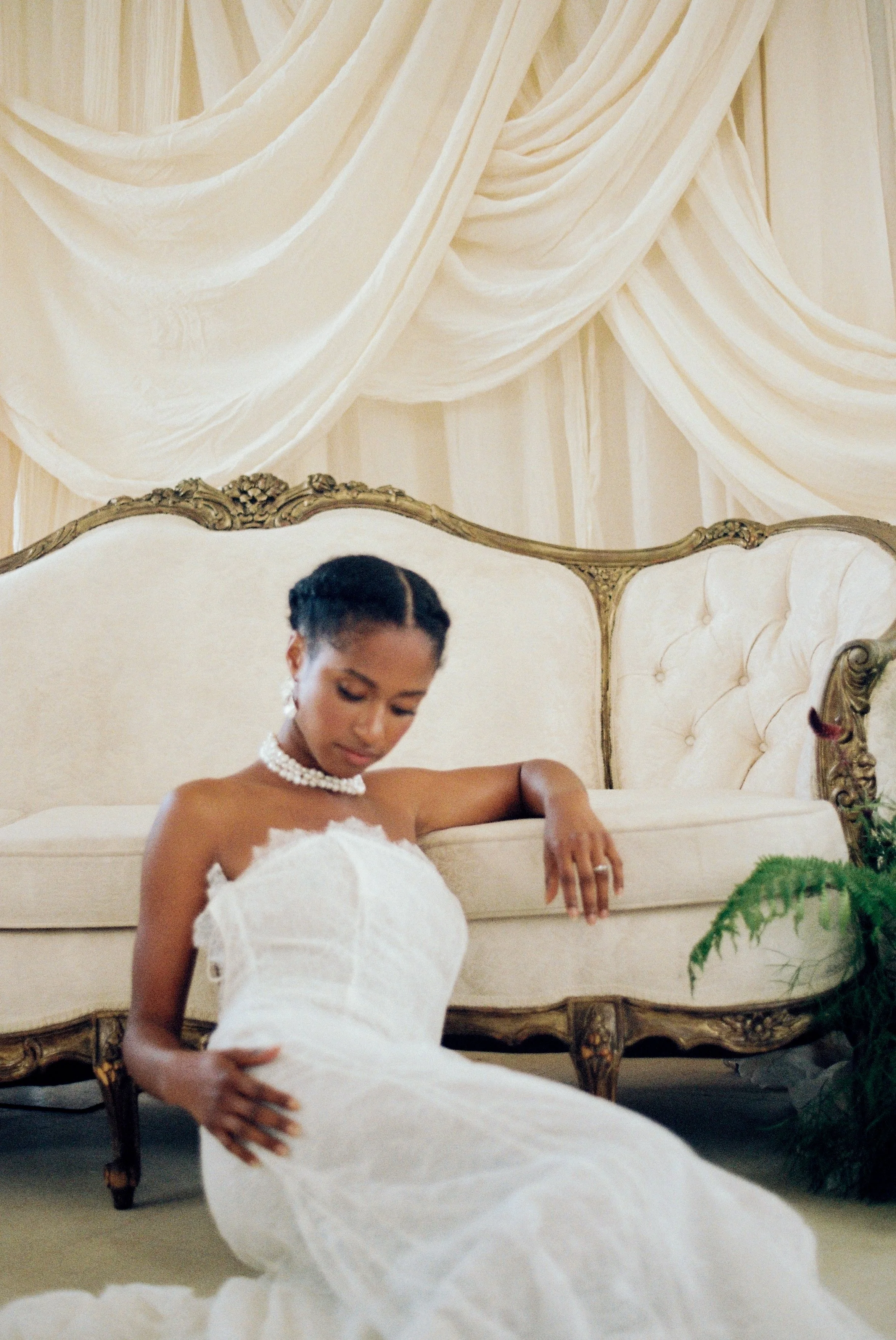 A bride in a white wedding dress with a pearl necklace, sitting on the floor in front of a vintage cream couch with ornate wooden trim, in a room with cream drapes on the wall.