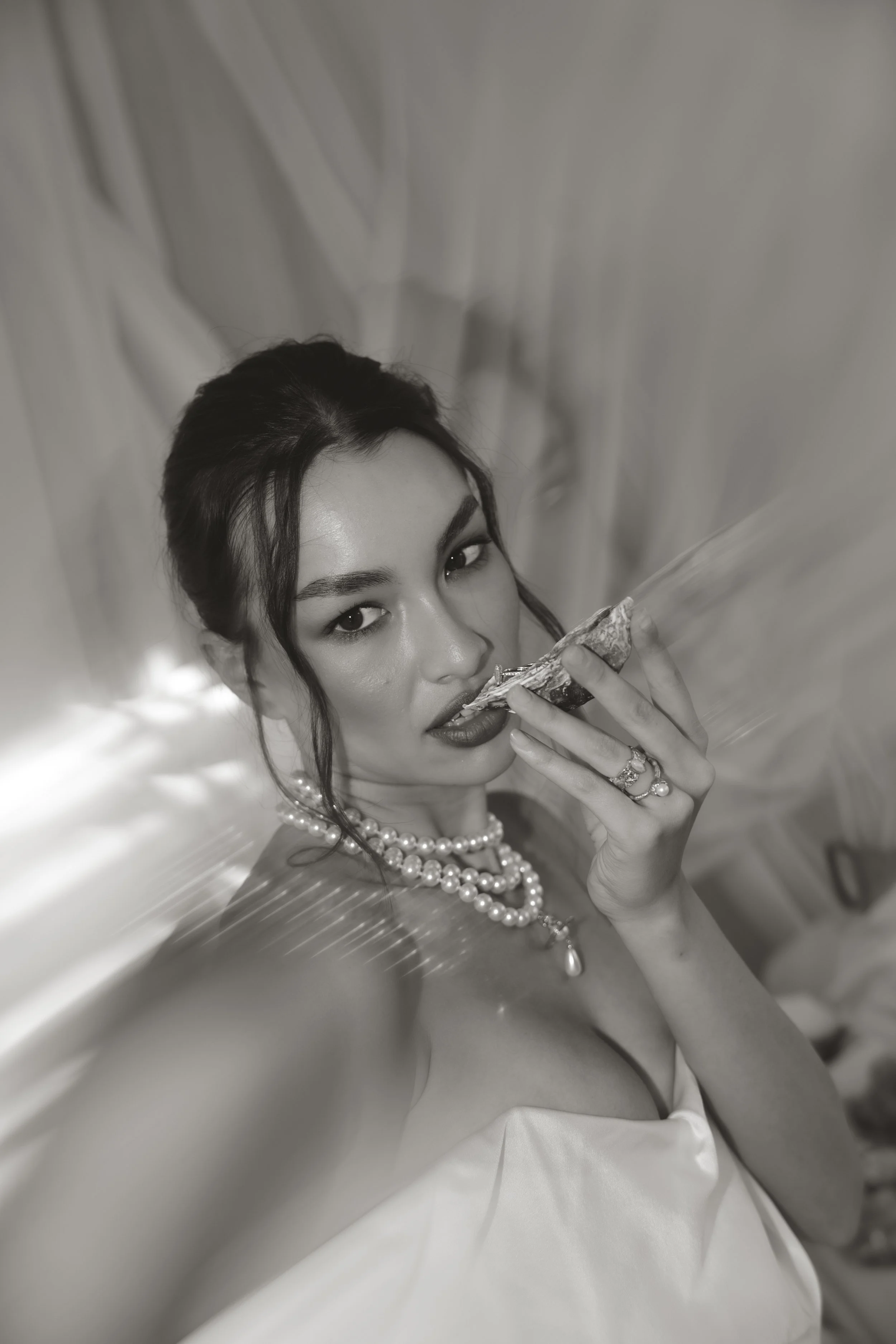 Editorial black and white portrait of a chic bride with an updo, wearing a luxury Vivienne Westwood wedding dress and pearl jewellery, holding an oyster against a curtain backdrop.