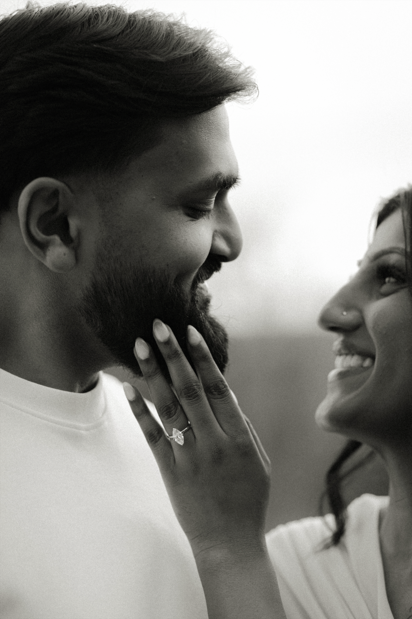 Close-up black and white engagement portrait of a romantic Asian couple smiling at each other, with the future bride touching her fiancé's face, showing her elegant engagement ring.