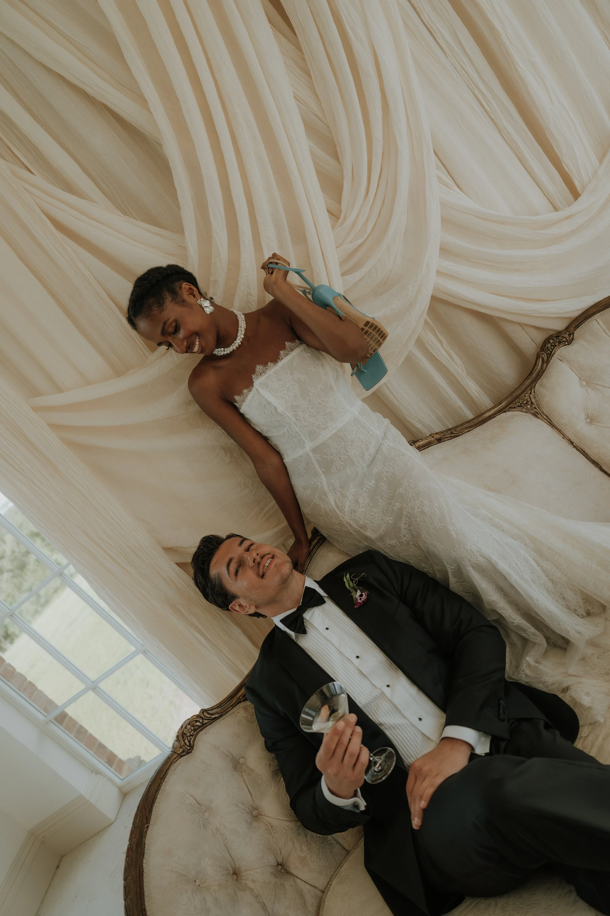 A playful, candid shot of a bride in a luxury Vera Wang wedding dress holding a hair straightener and a groom holding a wine glass, sitting together on a vintage sofa in front of large draped windows.