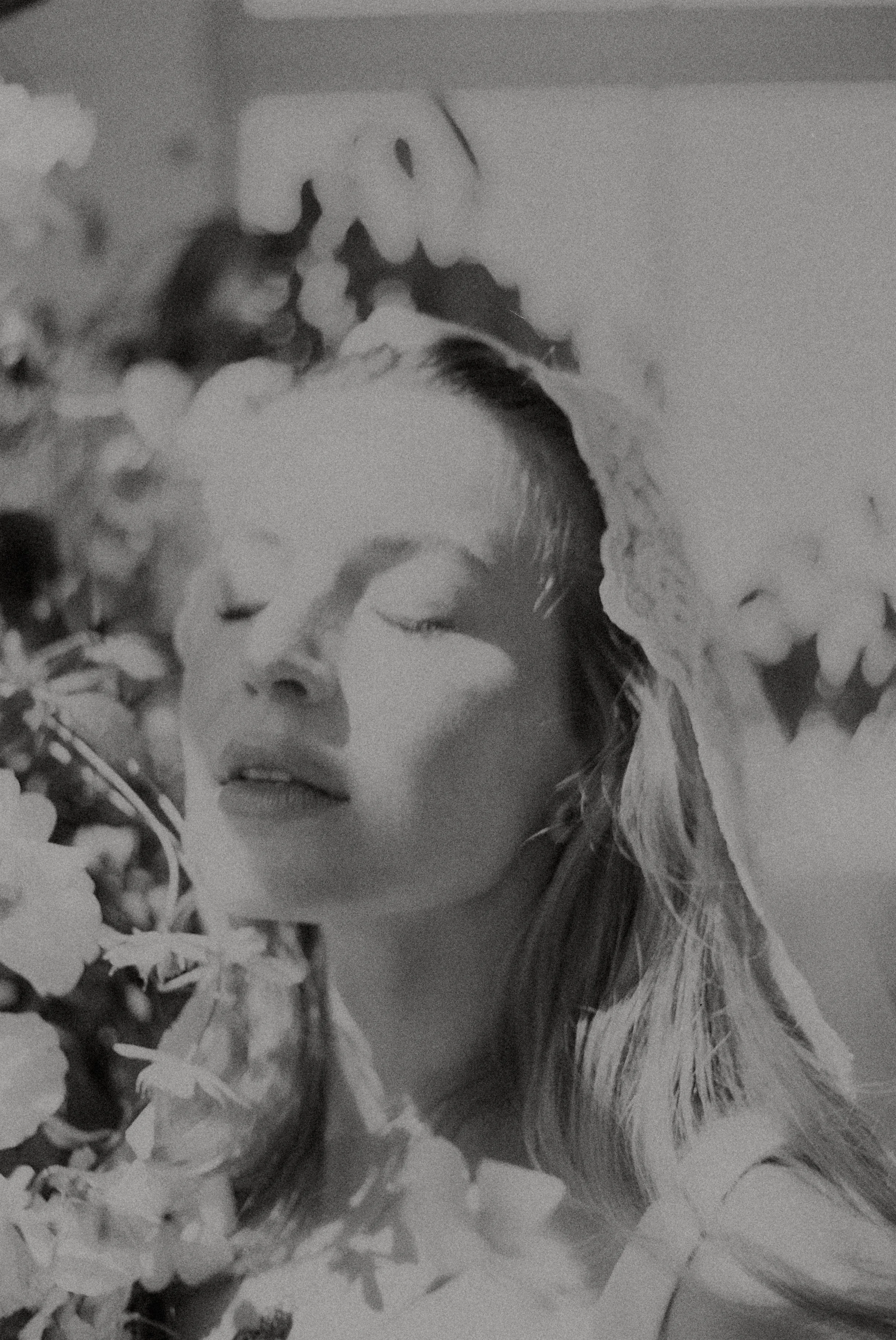 Black and white close-up photo of a bride's face with floral decorations around, featuring closed eyes and long hair.