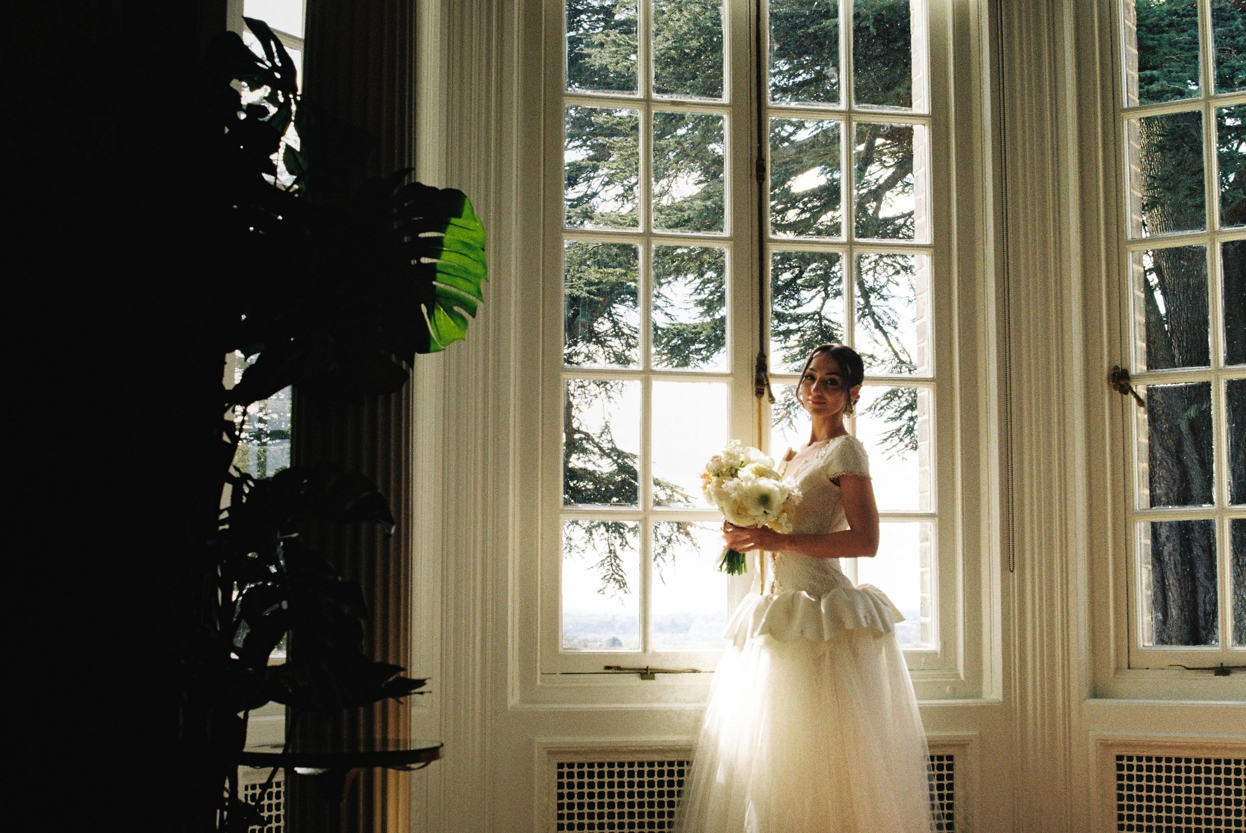 A bride in a white wedding dress holding a bouquet of white flowers, standing near a large window with trees outside, indoors with dark curtains and a potted plant in the foreground.