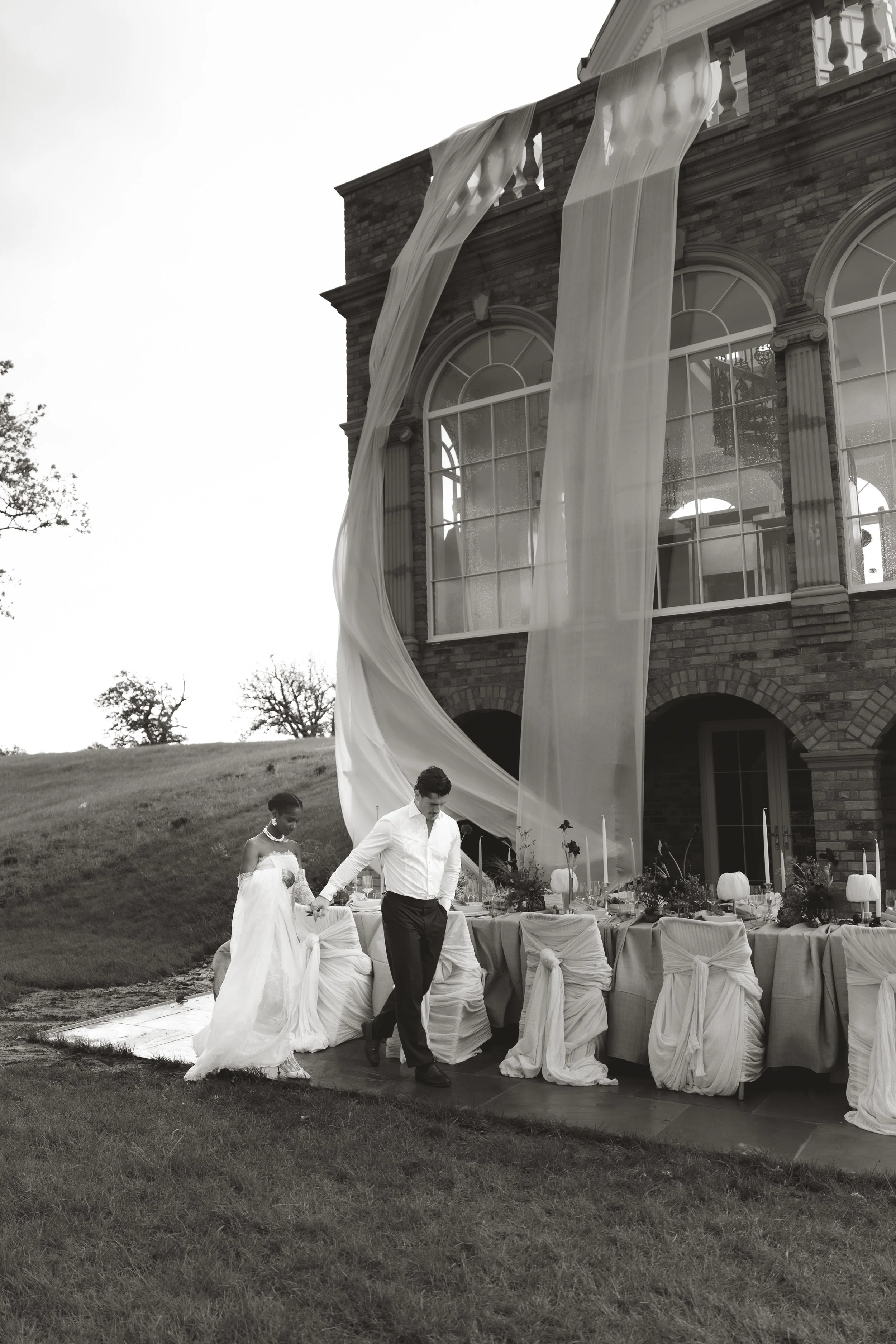 An elegant outdoor wedding reception scene featuring guests gathered in front of a historic brick building with large arched windows and dramatic, flowing white fabric installations.