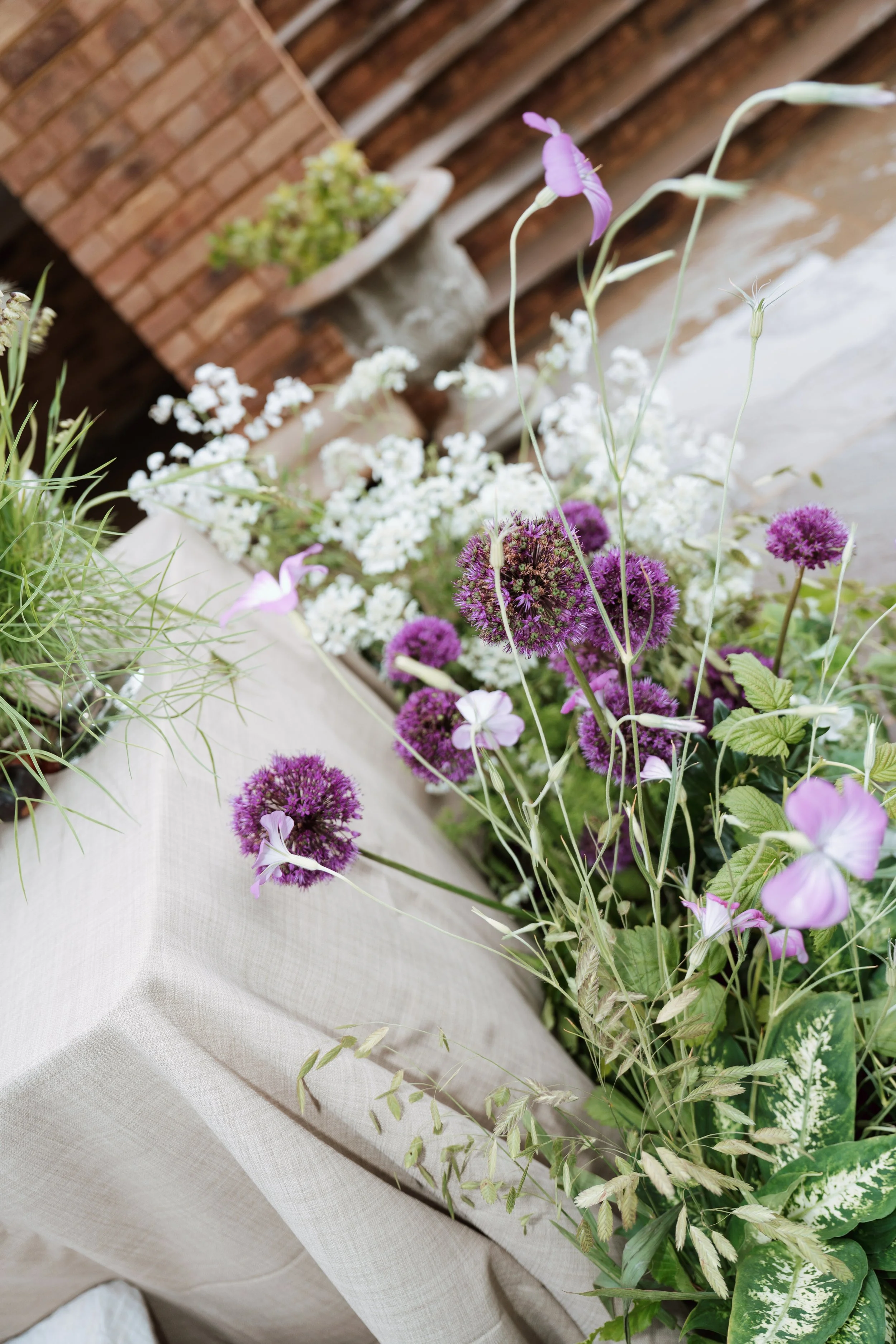 Close-up of a floral arrangement on a table with purple and white flowers, green foliage, and a brick wall in the background.