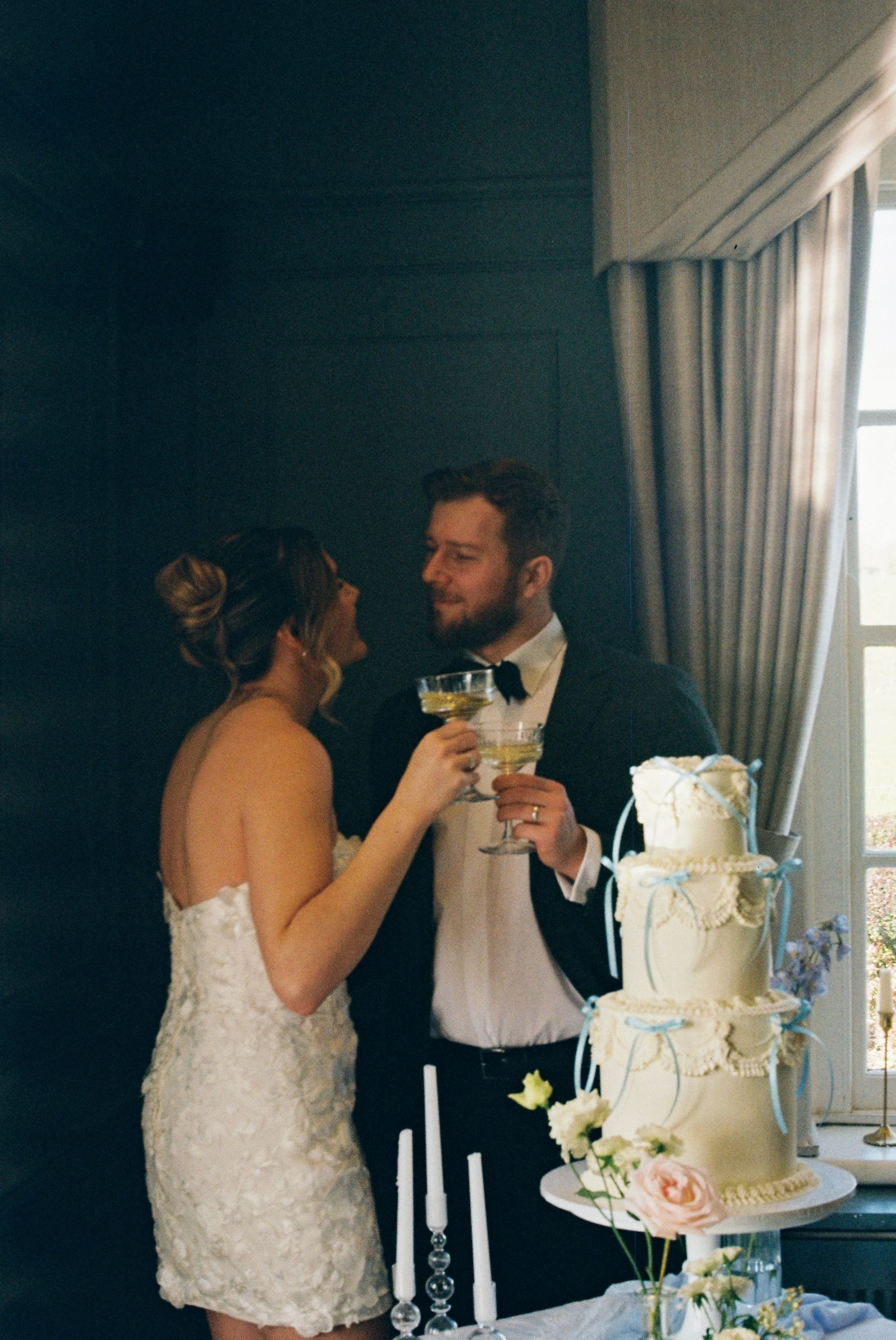 A bride and groom hold glasses of champagne, smiling at each other, beside a wedding cake decorated with blue ribbons and flowers at Downham Hall.