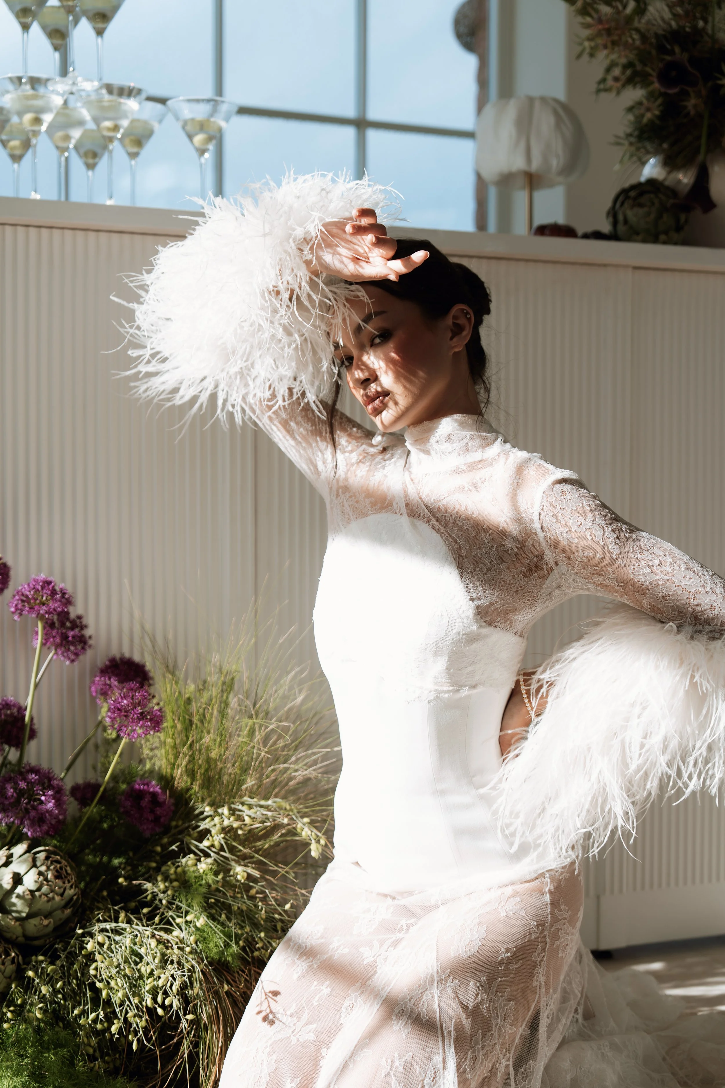 A woman in a white wedding dress with lace details and feathered accessories, raising her hand to her forehead, standing next to a floral arrangement with purple flowers in front of a large window.