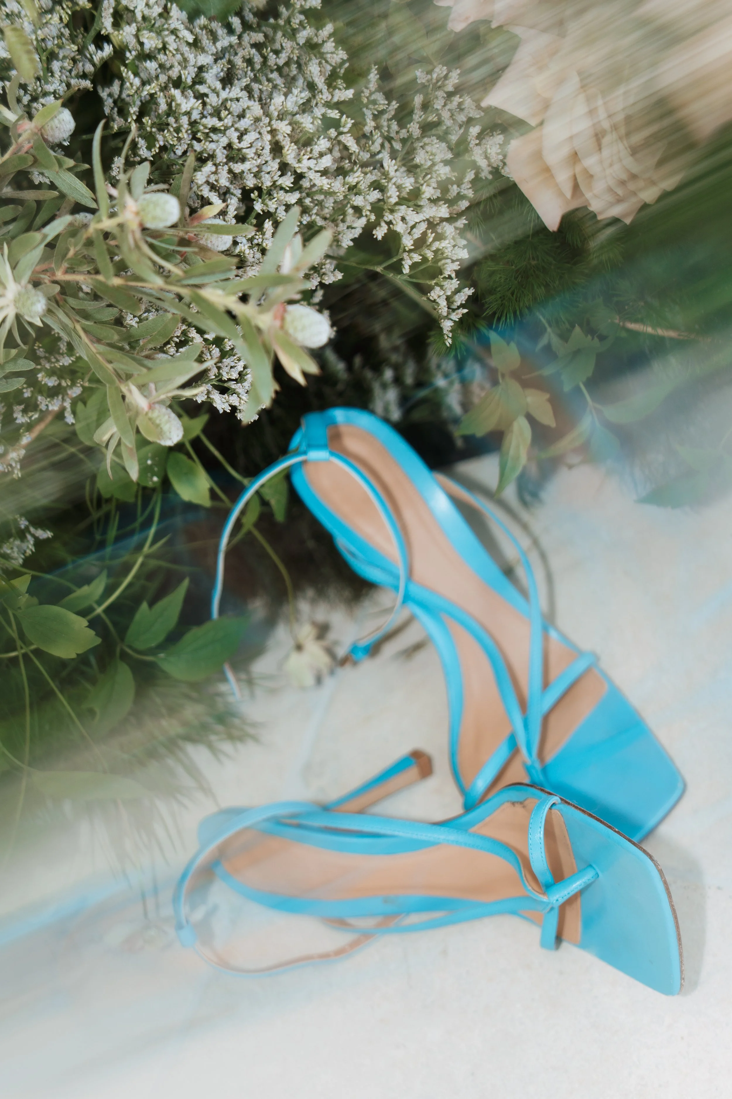 A luxury bridal detail shot of statement bright blue open-toe strappy high-heeled sandals placed on a neutral surface with green leafy plants.