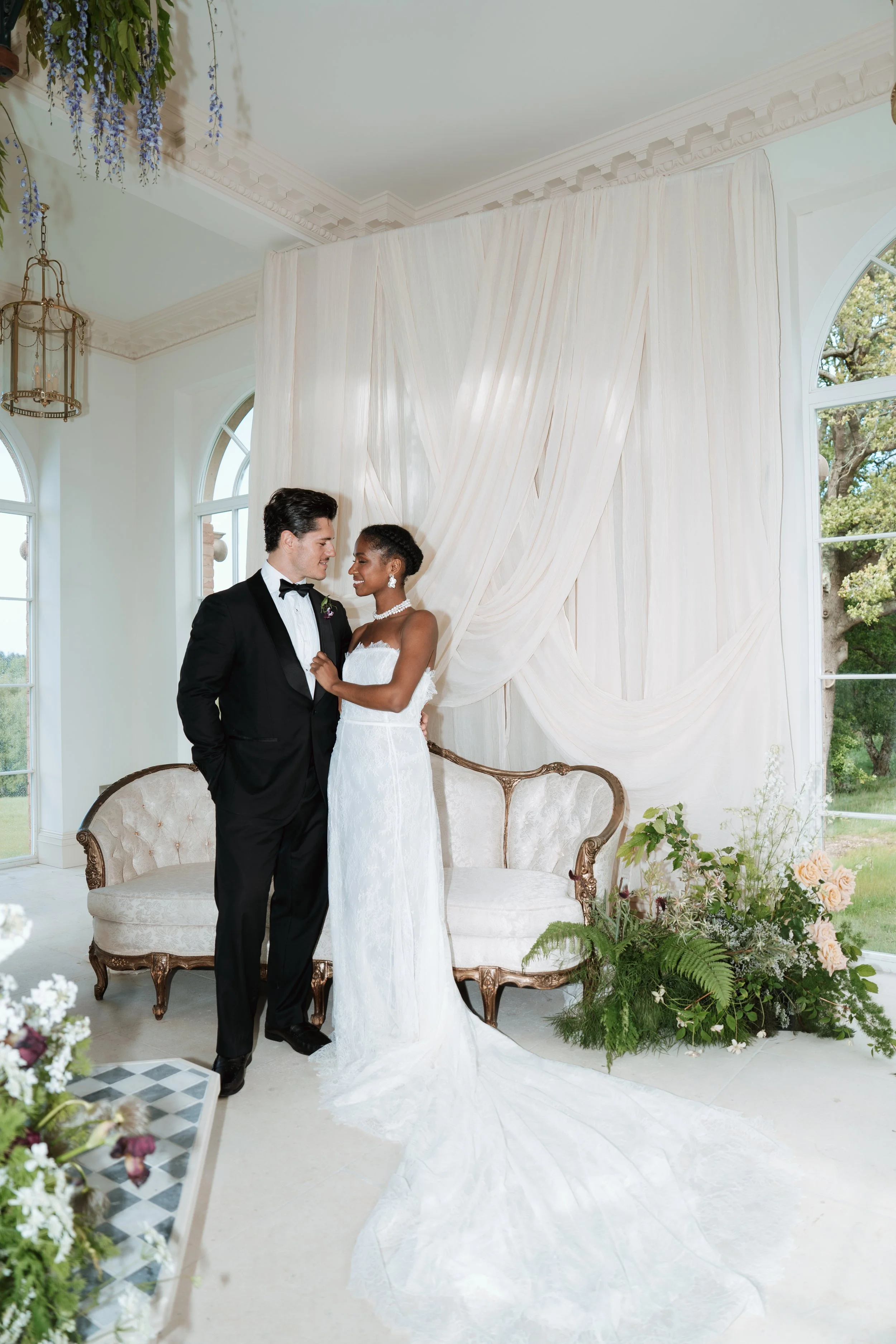 A romantic indoor wedding scene featuring a groom in a black tuxedo and a bride in a stunning Vera Wang wedding gown, smiling at each other in front of elegant draped white fabric and floral arrangements.