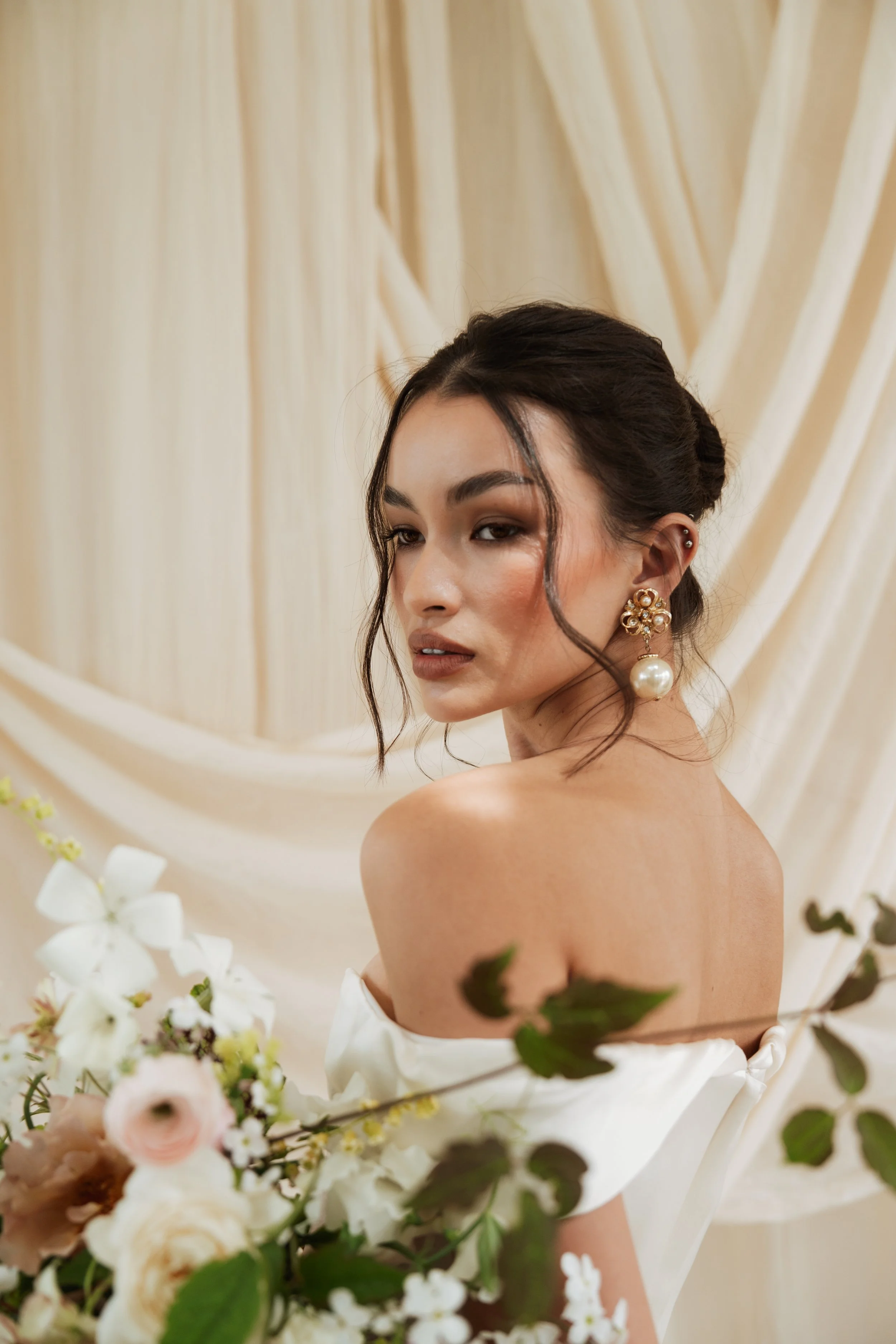 Editorial bridal portrait of a woman with an elegant bun updo wearing a strapless Vivienne Westwood wedding dress, holding a bouquet of white and pink flowers against a draped cream curtain.