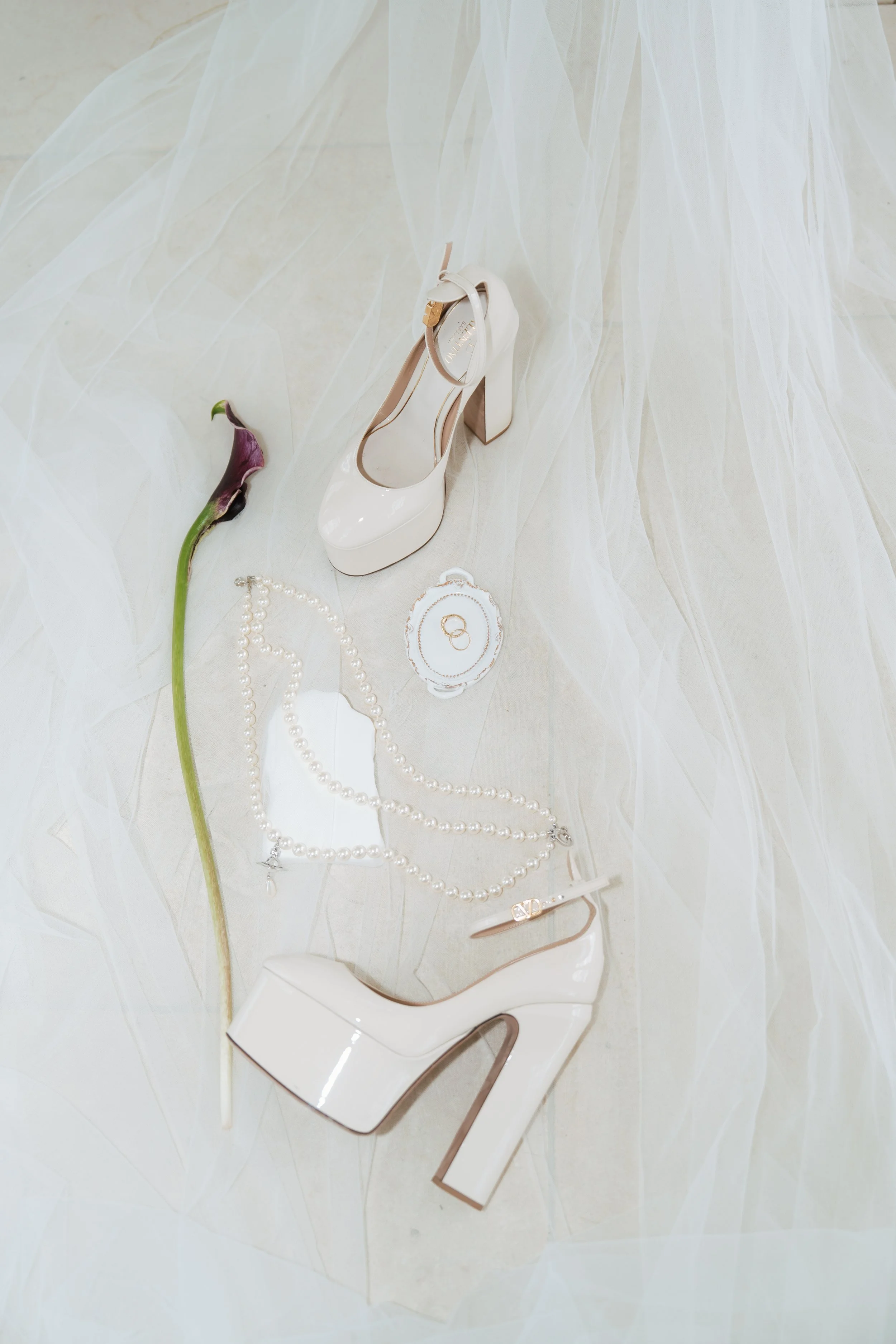 A luxury wedding flat lay style display featuring white Valentino bridal shoes with an elegant bow ankle strap, styled with a pearl necklace and sheer fabric.